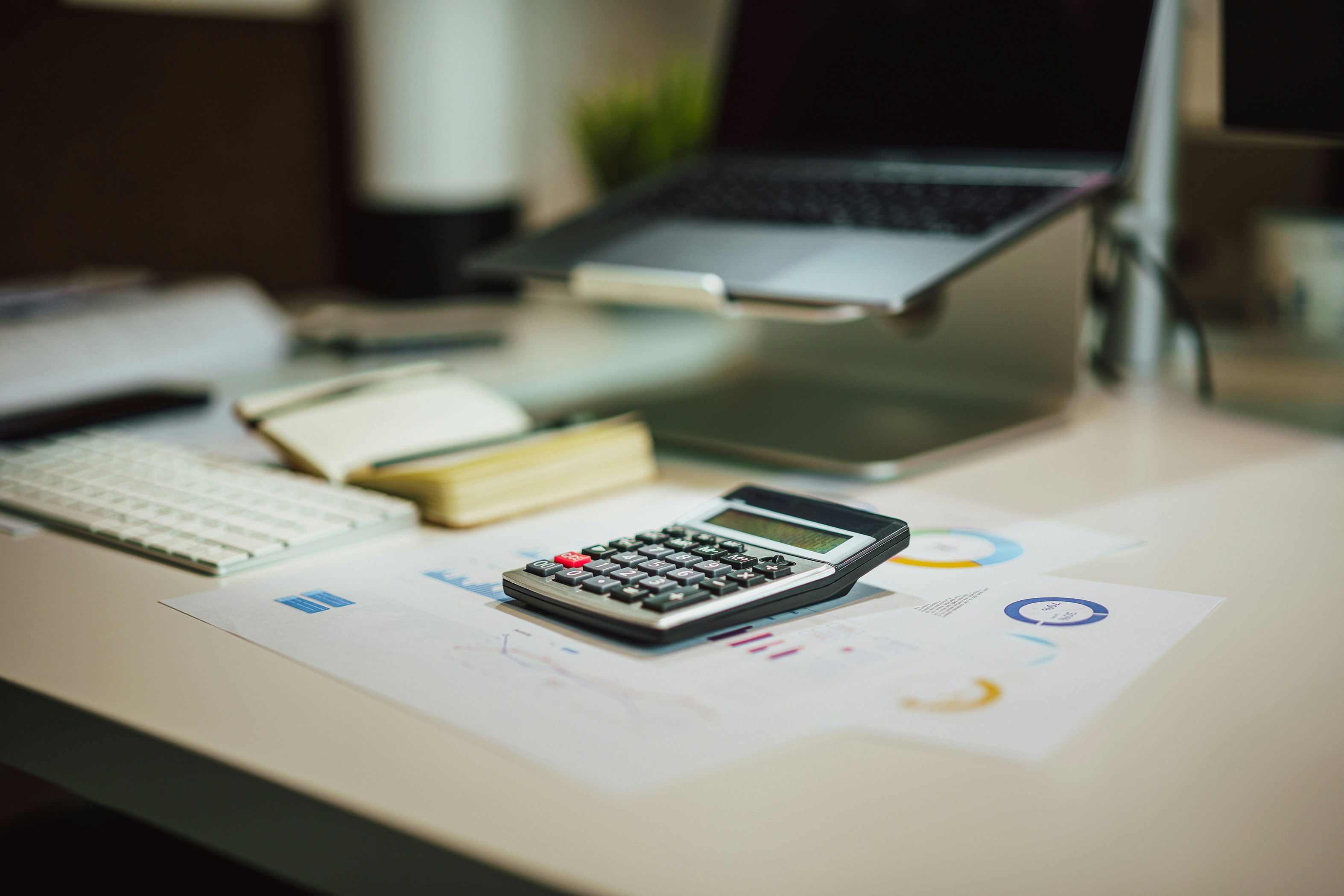 Calculator on financial documents alongside a laptop, keyboard and notebook on an office desk.