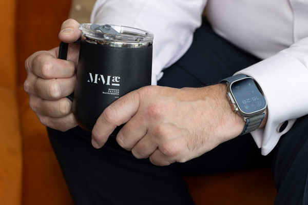 Close‑up of a person in business attire holding a black MFMac branded travel mug, wearing a smartwatch.