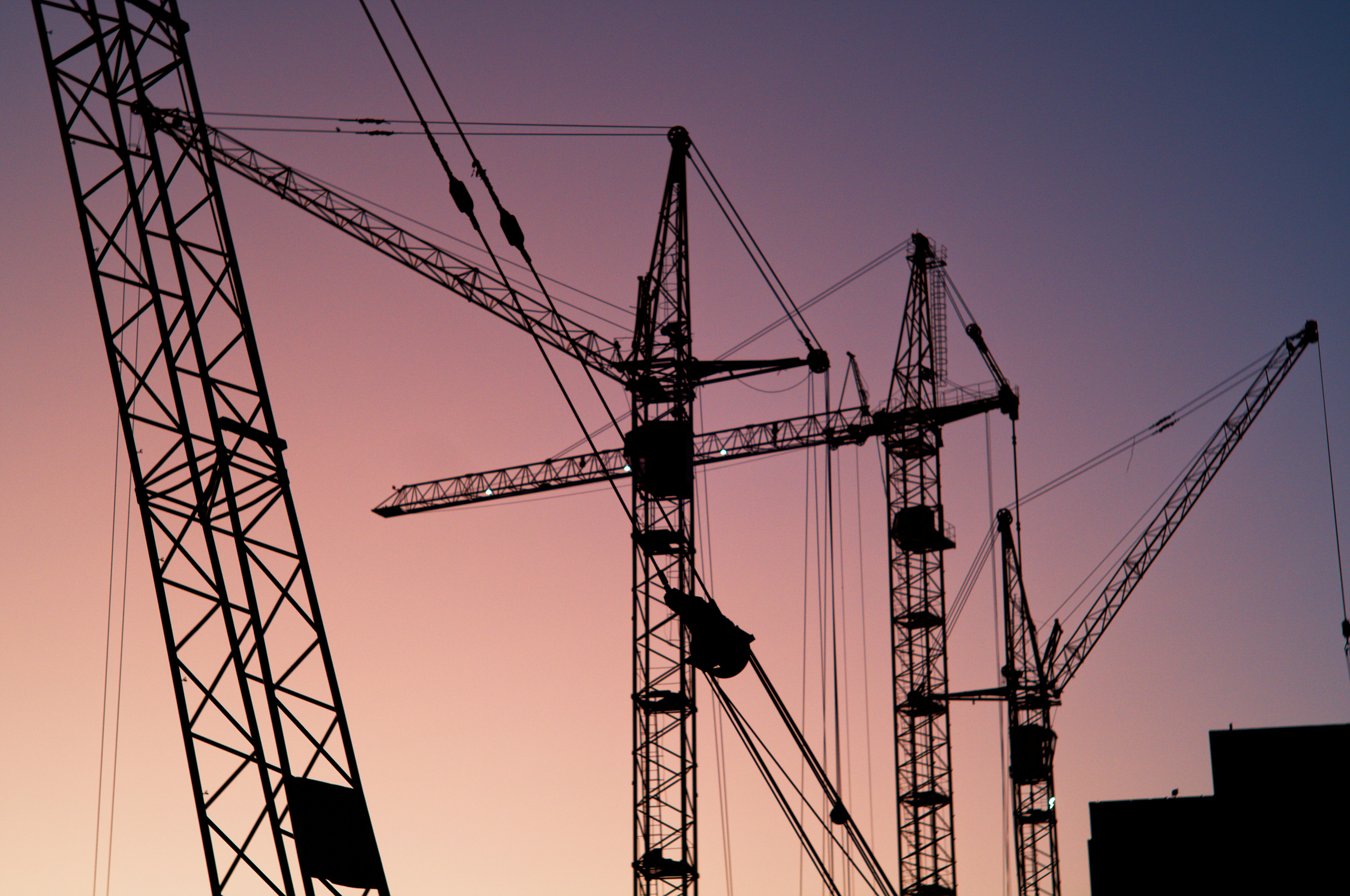 Construction cranes silhouetted against a pink and purple evening sky.