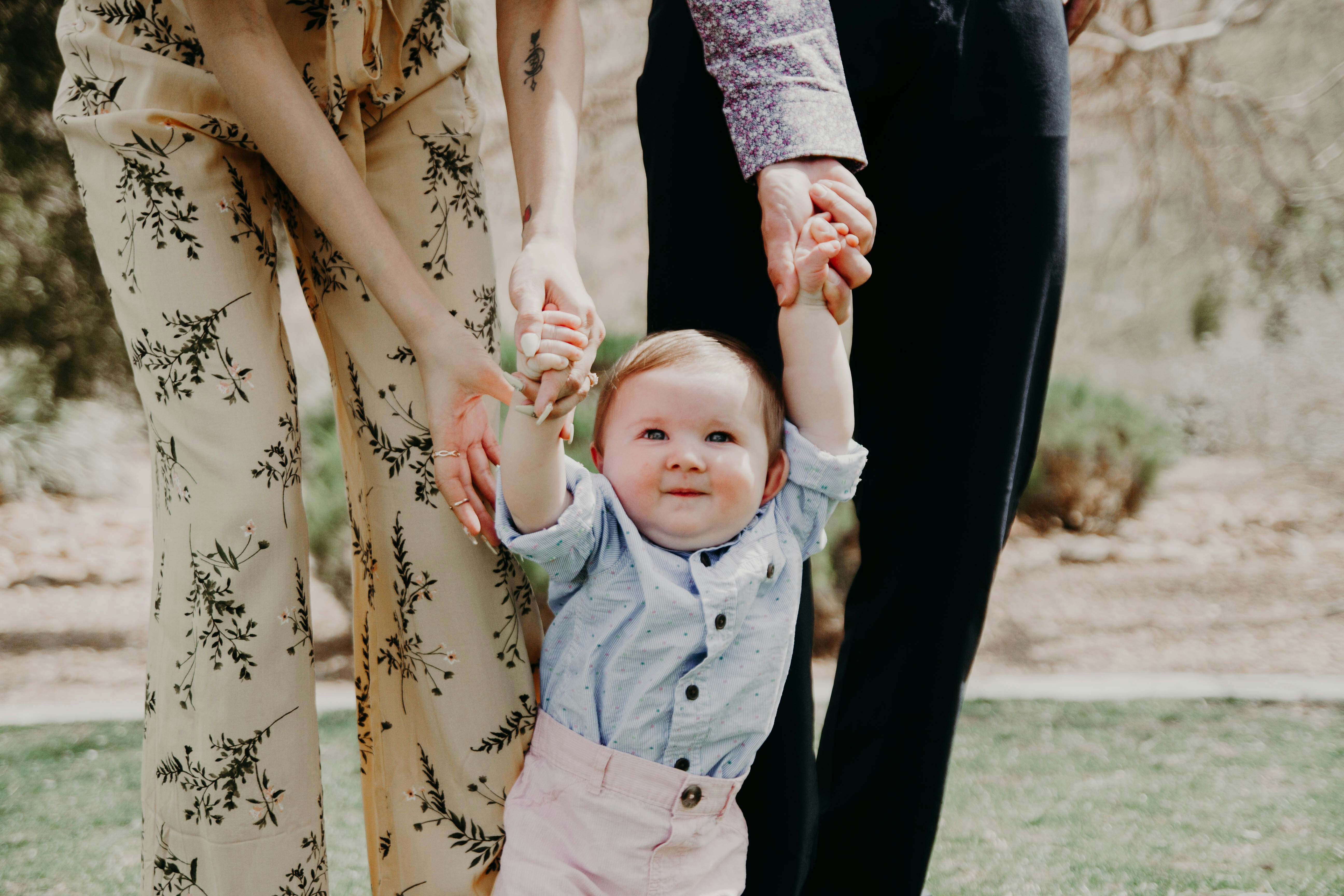 A young child being supported while standing outdoors, holding the hands of two adults.