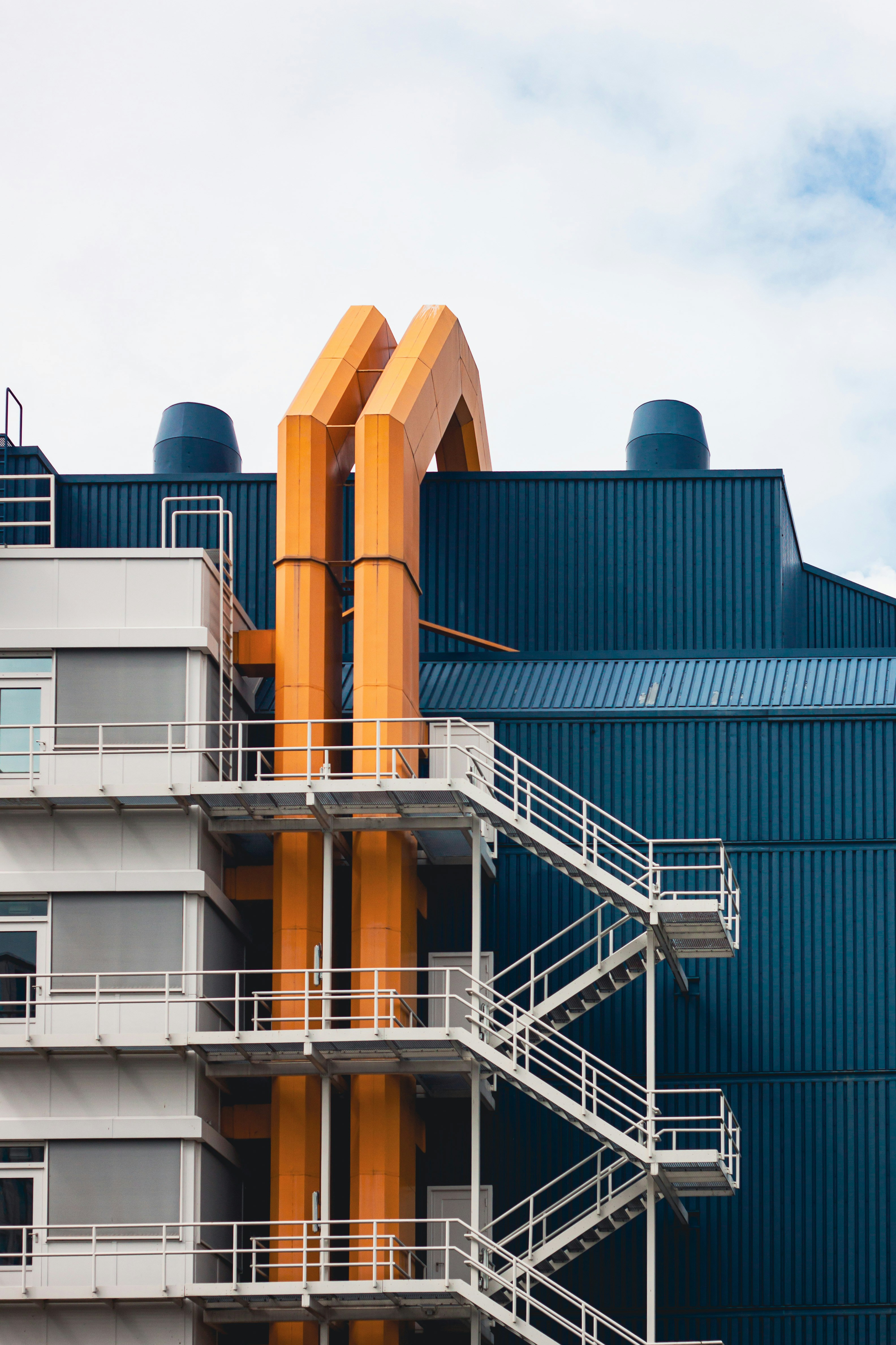 Close-up view of a contemporary industrial building featuring blue corrugated metal panels, bright orange structural pipes, and white metal staircases on the exterior, set against a partly cloudy sky.