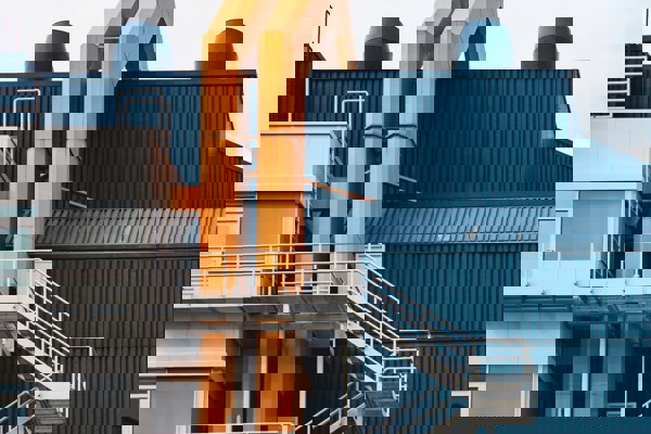 Close-up view of a contemporary industrial building featuring blue corrugated metal panels, bright orange structural pipes, and white metal staircases on the exterior, set against a partly cloudy sky.