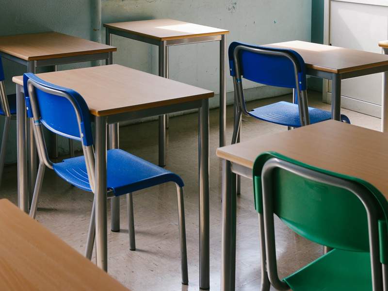 Empty classroom with several wooden desks and colourful plastic chairs, including blue and green ones, arranged in rows on a tiled floor. The room has light-coloured walls and natural light coming from a window on the right.