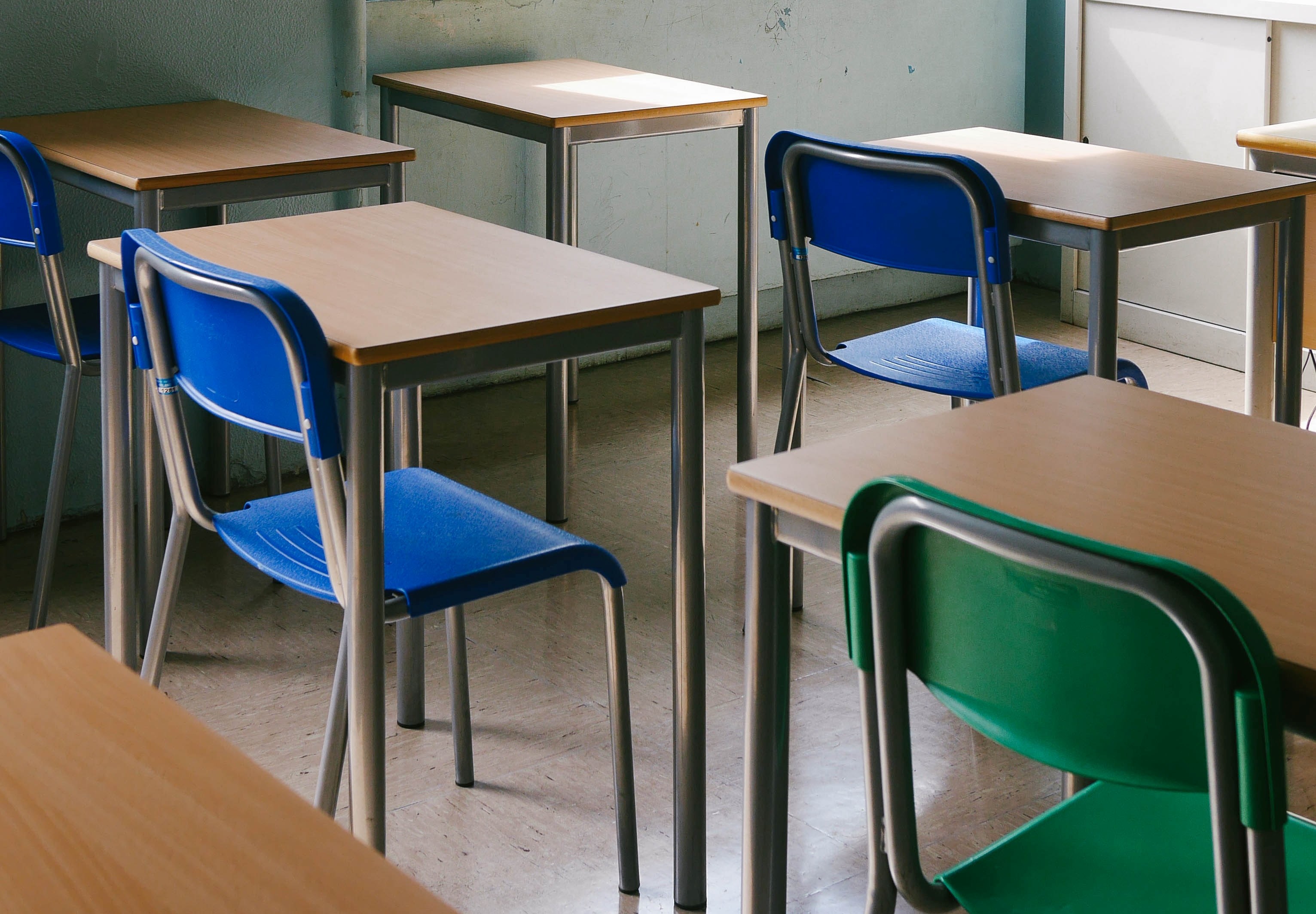 Empty classroom with several wooden desks and colourful plastic chairs, including blue and green ones, arranged in rows on a tiled floor. The room has light-coloured walls and natural light coming from a window on the right.
