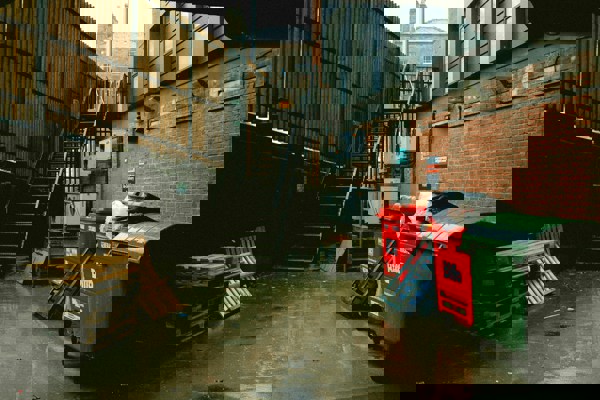 Outdoor service area with bins, wooden pallets and a metal staircase beside brick buildings on a wet day.