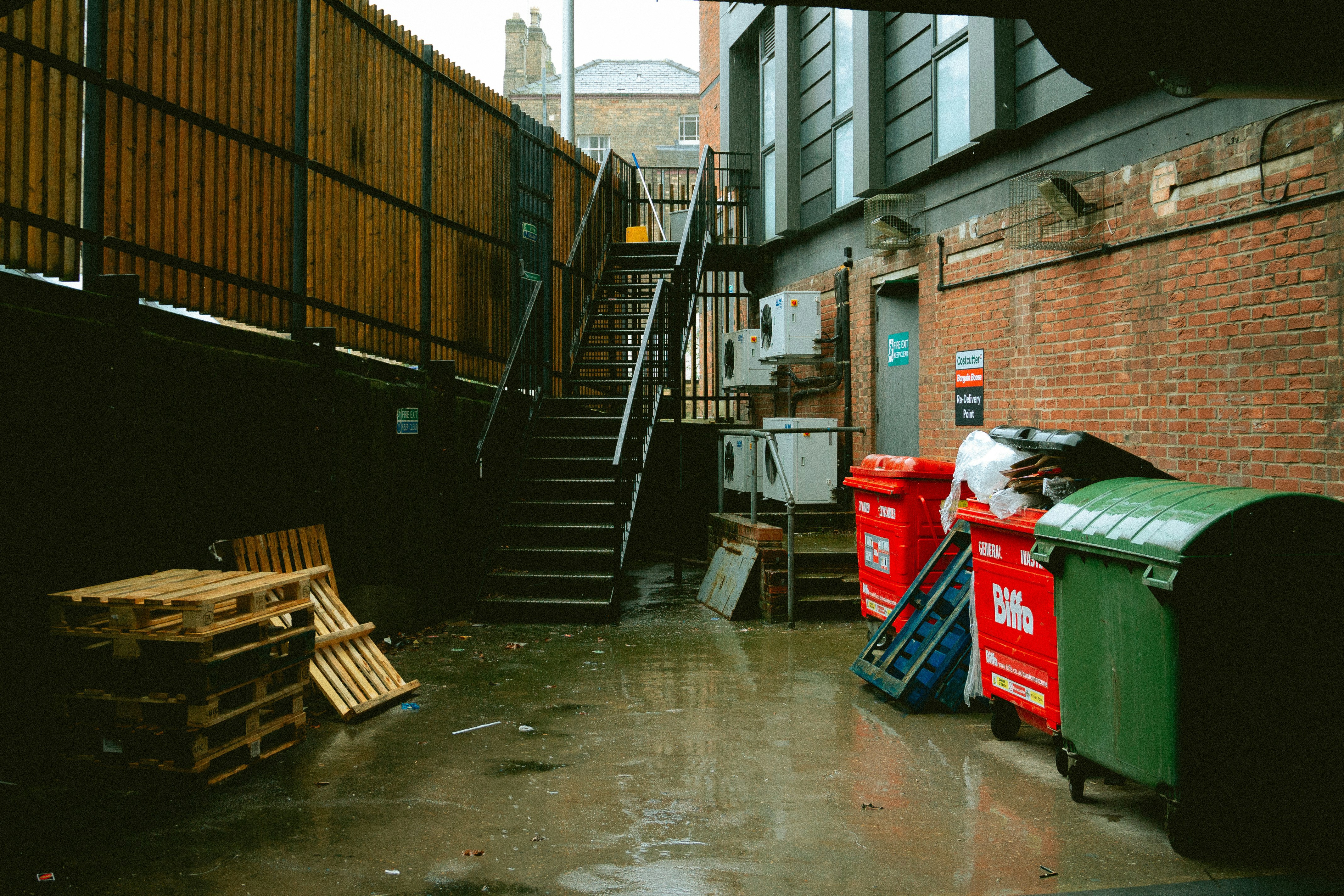 Outdoor service area with bins, wooden pallets and a metal staircase beside brick buildings on a wet day.