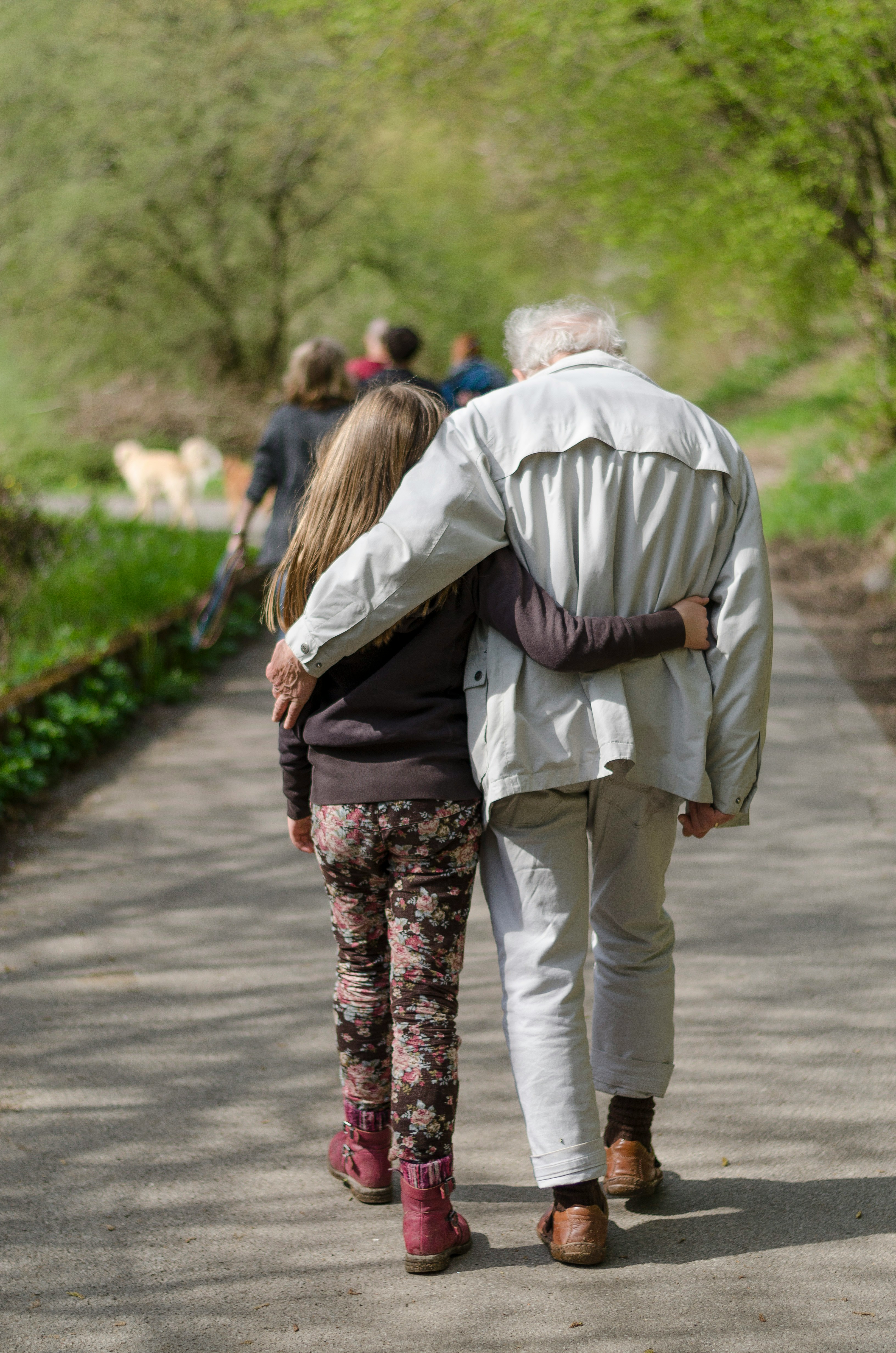 Two people walking together along a path in a park, with one person’s arm around the other, seen from behind.