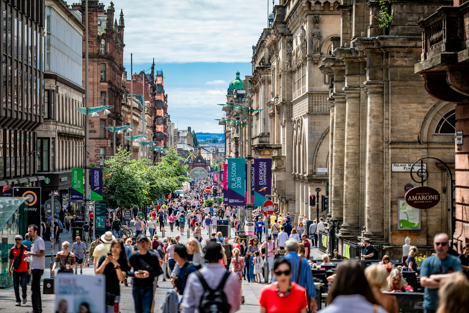 Busy pedestrianised street in central Glasgow lined with historic stone buildings and modern shopfronts, with large crowds of people walking along the avenue beneath event banners and a bright, partly cloudy sky.