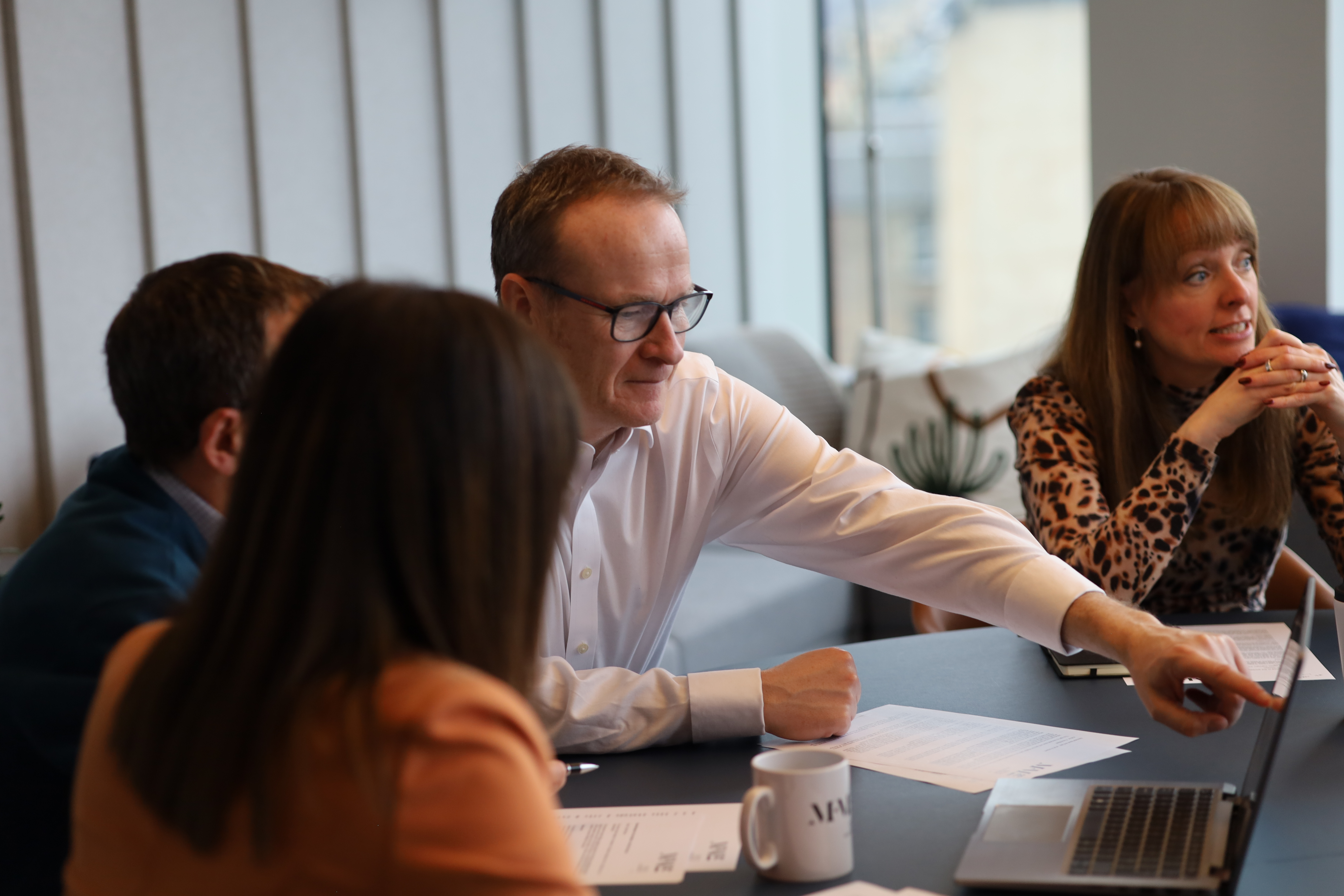 People gathered around a meeting table in a modern office, reviewing documents and discussing information displayed on a laptop during a team meeting.