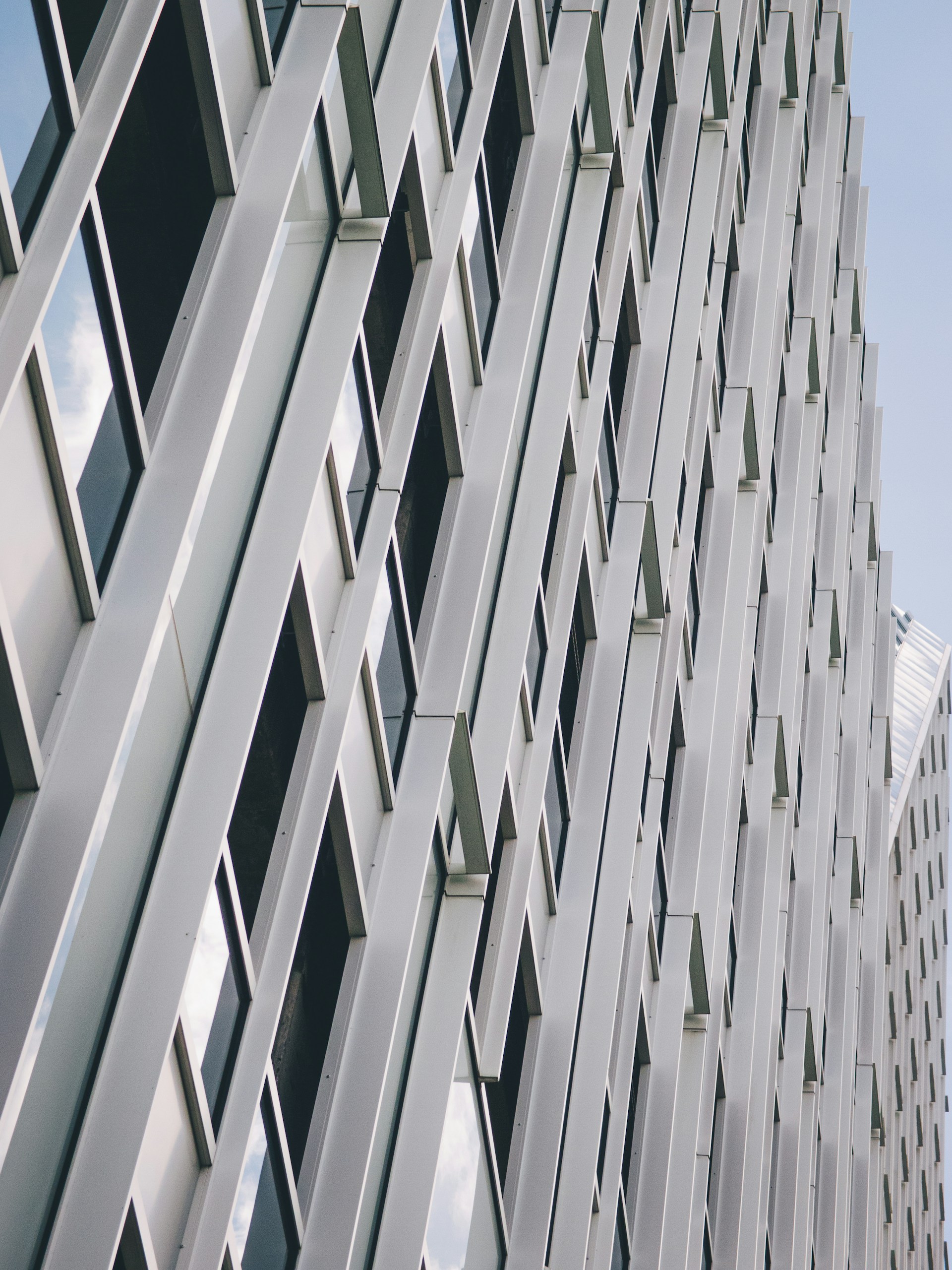 Close-up view of a modern building façade with repeating vertical and diagonal metal panels forming a geometric pattern against a pale sky.