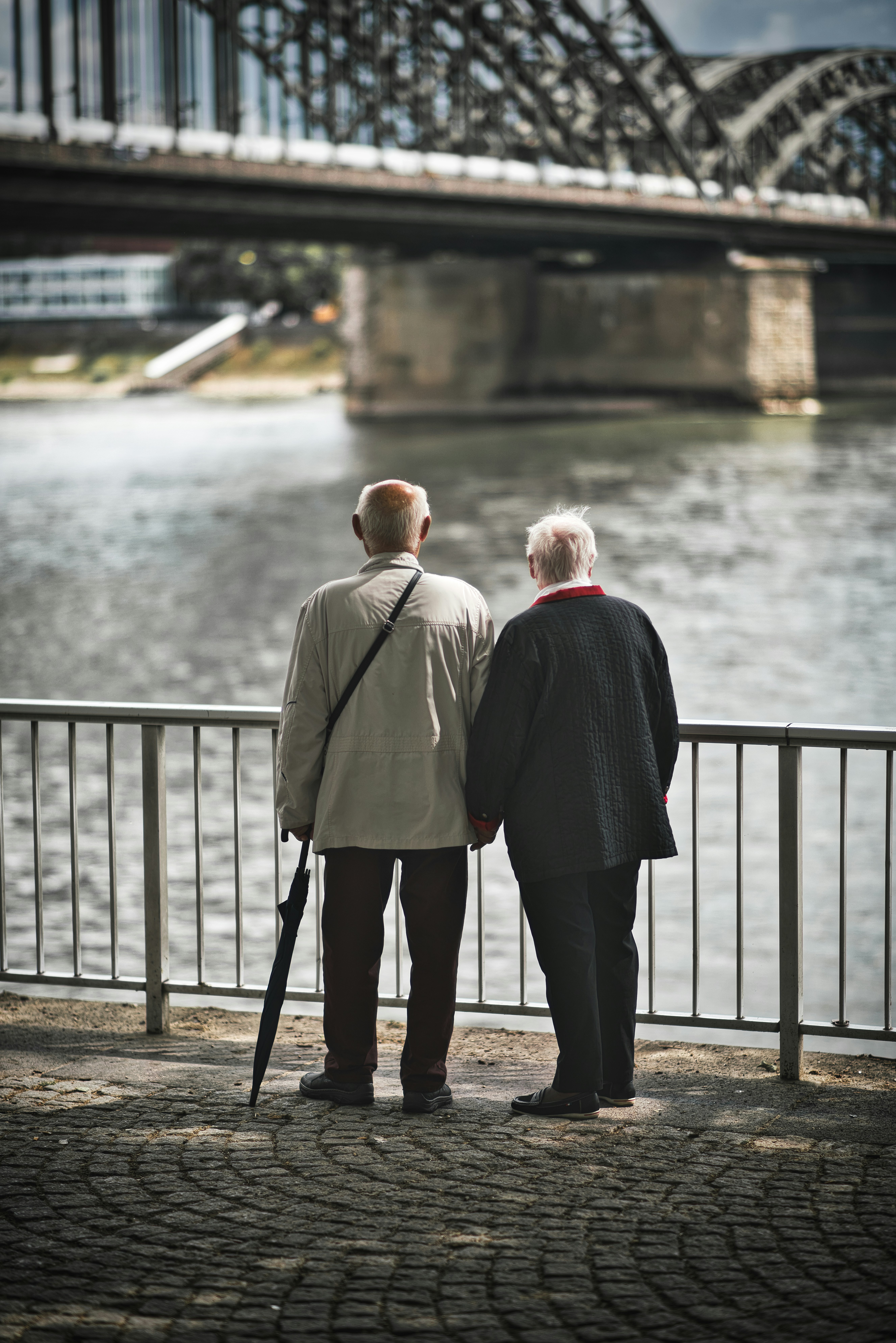 Two older people standing together by a riverside railing, looking out over the water in an urban setting.