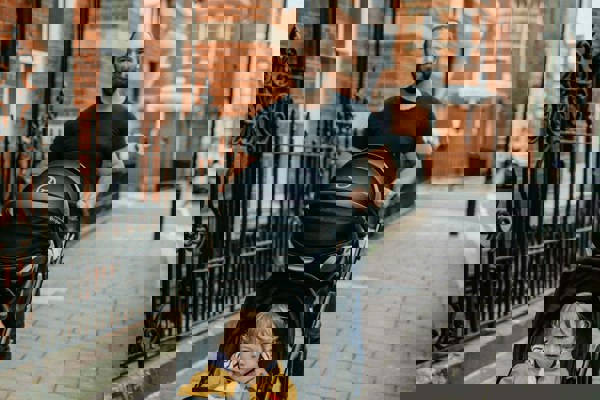 An adult pushing a pram with a young child along a pavement on a residential street lined with brick buildings and railings.