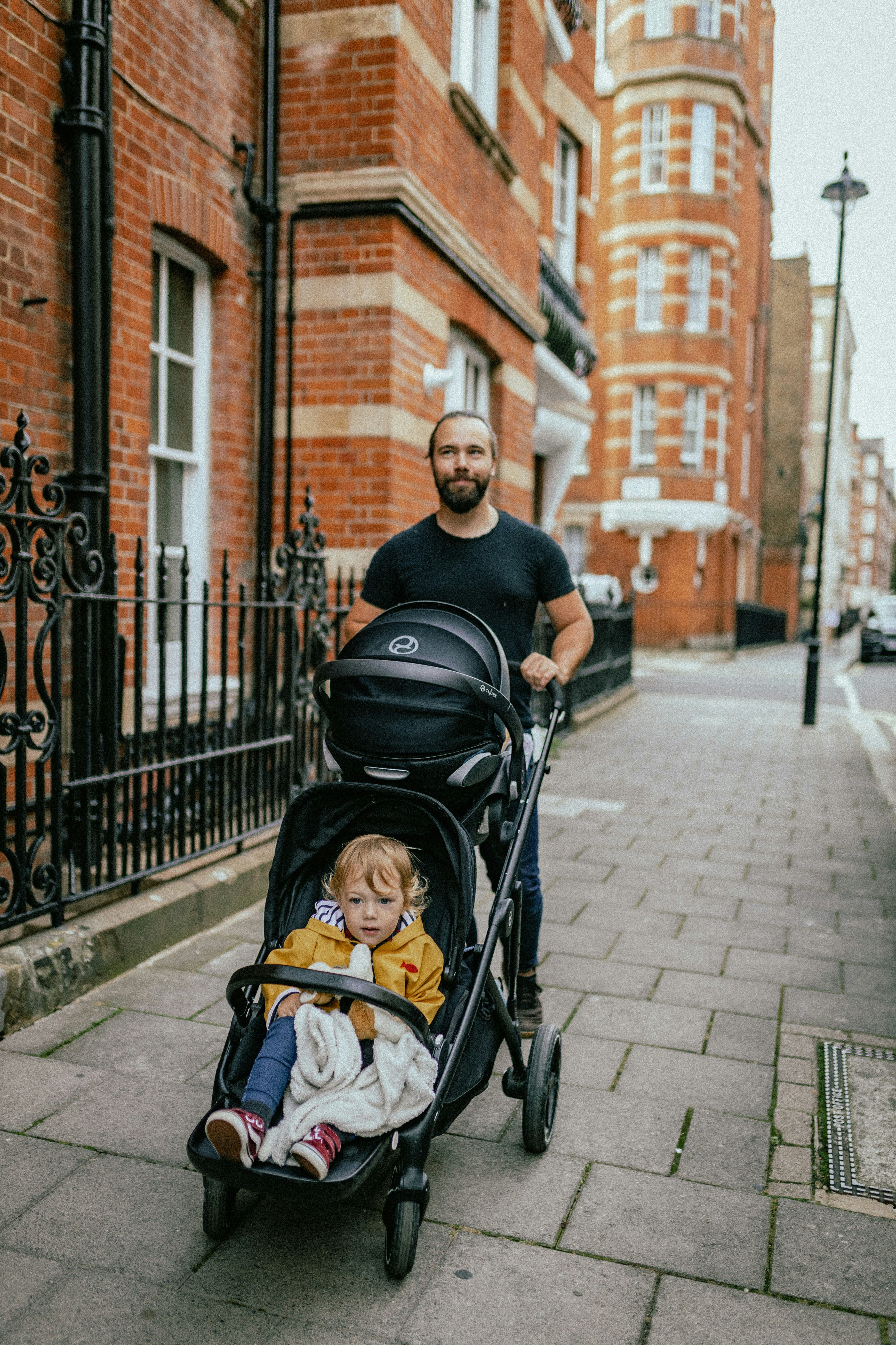 An adult pushing a pram with a young child along a pavement on a residential street lined with brick buildings and railings.