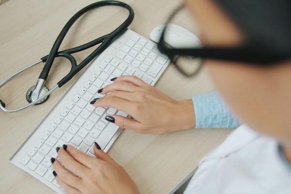 Healthcare professional using a computer, with a stethoscope resting on a desk beside a keyboard.