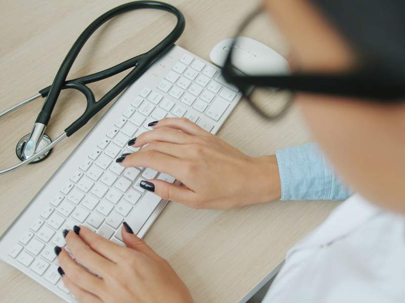 Healthcare professional using a computer, with a stethoscope resting on a desk beside a keyboard.