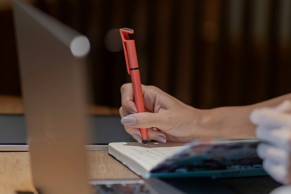 A person taking handwritten notes in a notebook beside an open laptop, suggesting human review and analysis.