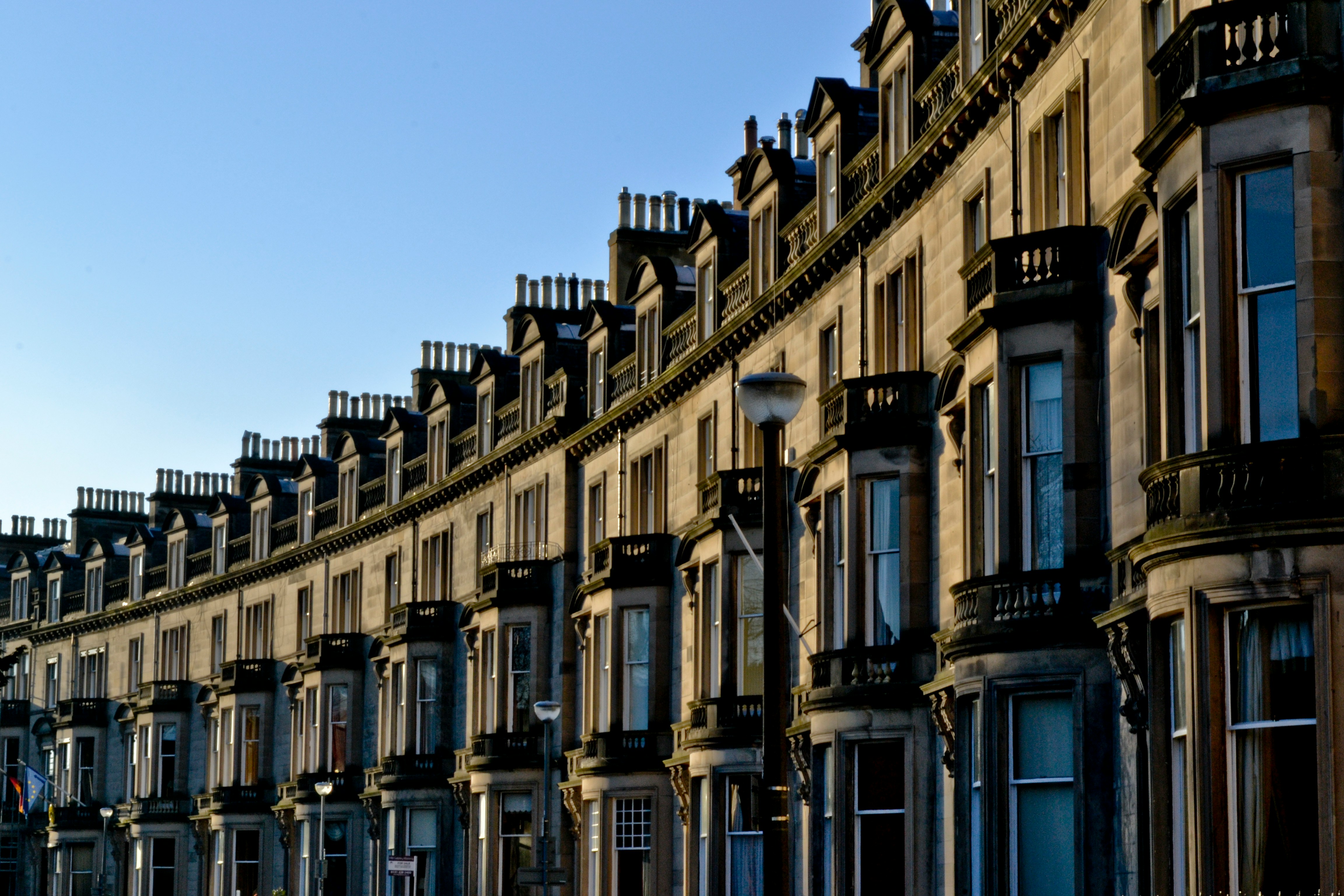 Curved terrace of traditional sandstone Edinburgh townhouses with bay windows, balconies and chimneys under a clear blue sky, representing life in the UK for visa applicants
