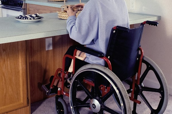 Person using a wheelchair seated at a kitchen counter, with cupboards, appliances and baskets visible in a domestic setting.
