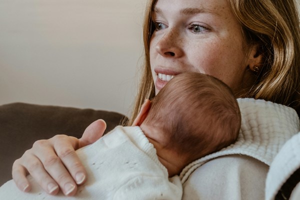 Person seated on a sofa holding a small baby resting against their shoulder, both wearing light-coloured clothing in a softly lit home setting.