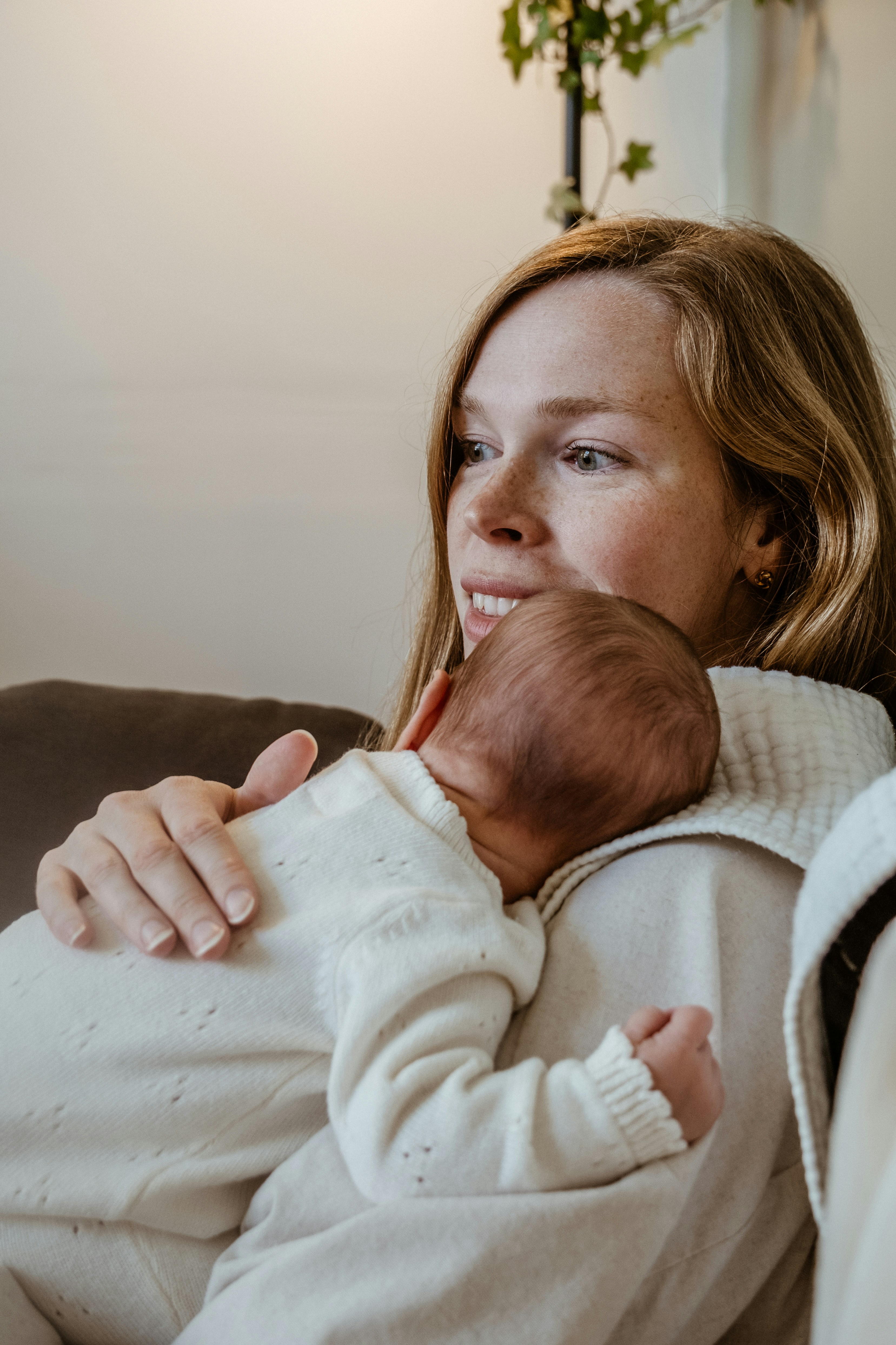 Person seated on a sofa holding a small baby resting against their shoulder, both wearing light-coloured clothing in a softly lit home setting.