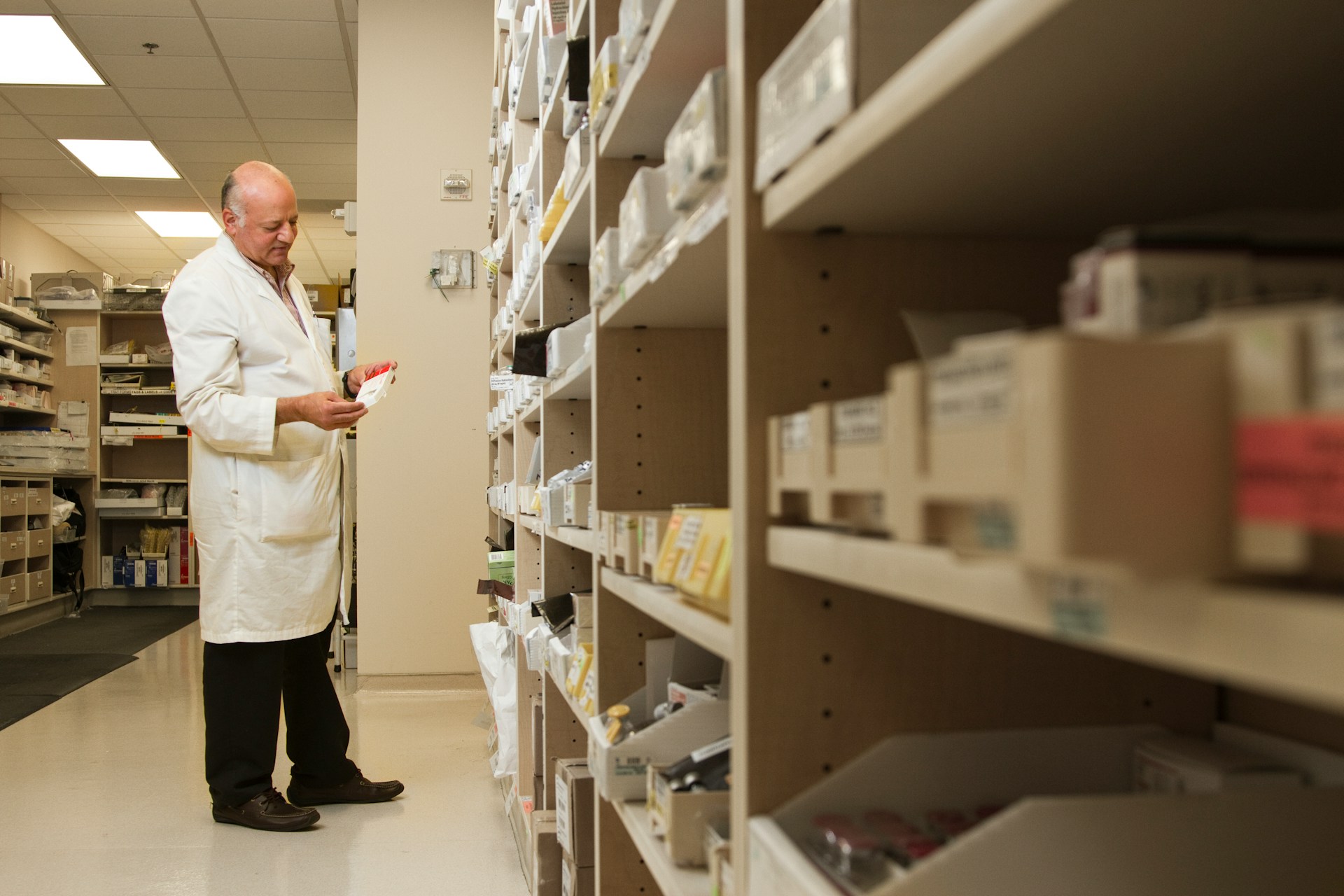 Pharmacist reviewing a medication box in a pharmacy dispensary, standing beside shelves stocked with labelled prescriptions.