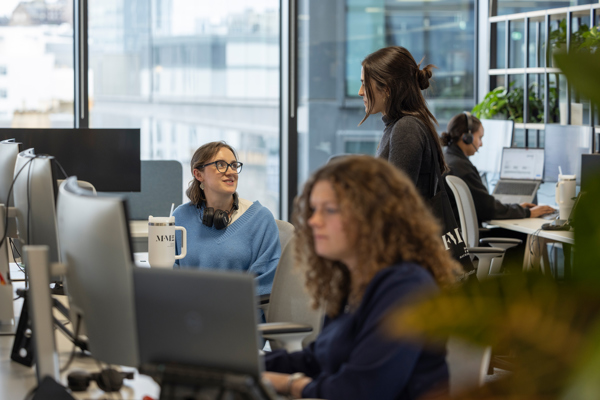 Office environment with people working at desks, using computers and headsets, and speaking with colleagues in an open‑plan workspace.