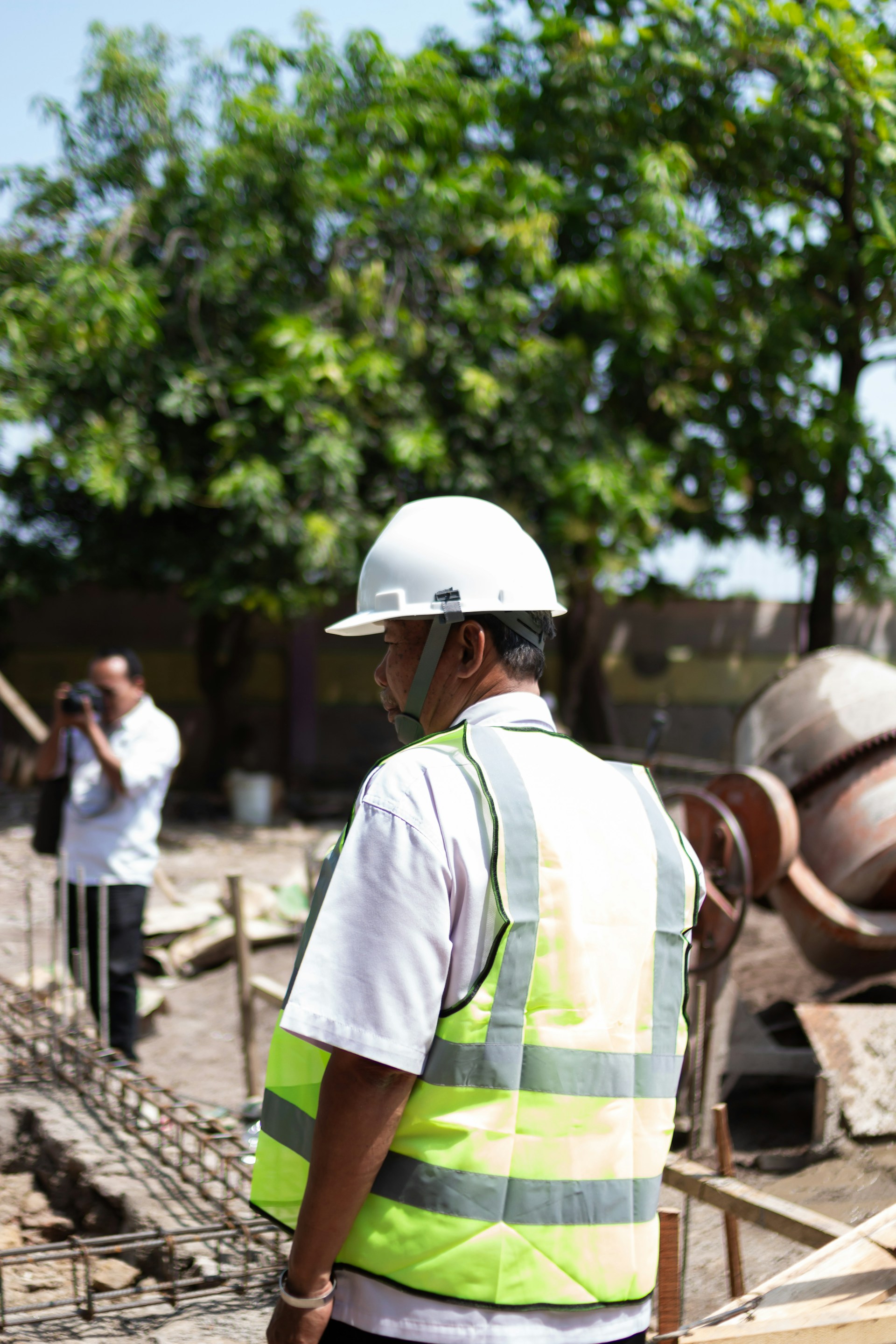 A construction worker wearing a hard hat and high-visibility vest standing on an active construction site beside reinforced foundations and concrete mixing equipment.