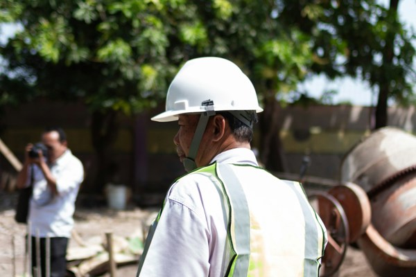 A construction worker wearing a hard hat and high-visibility vest standing on an active construction site beside reinforced foundations and concrete mixing equipment.