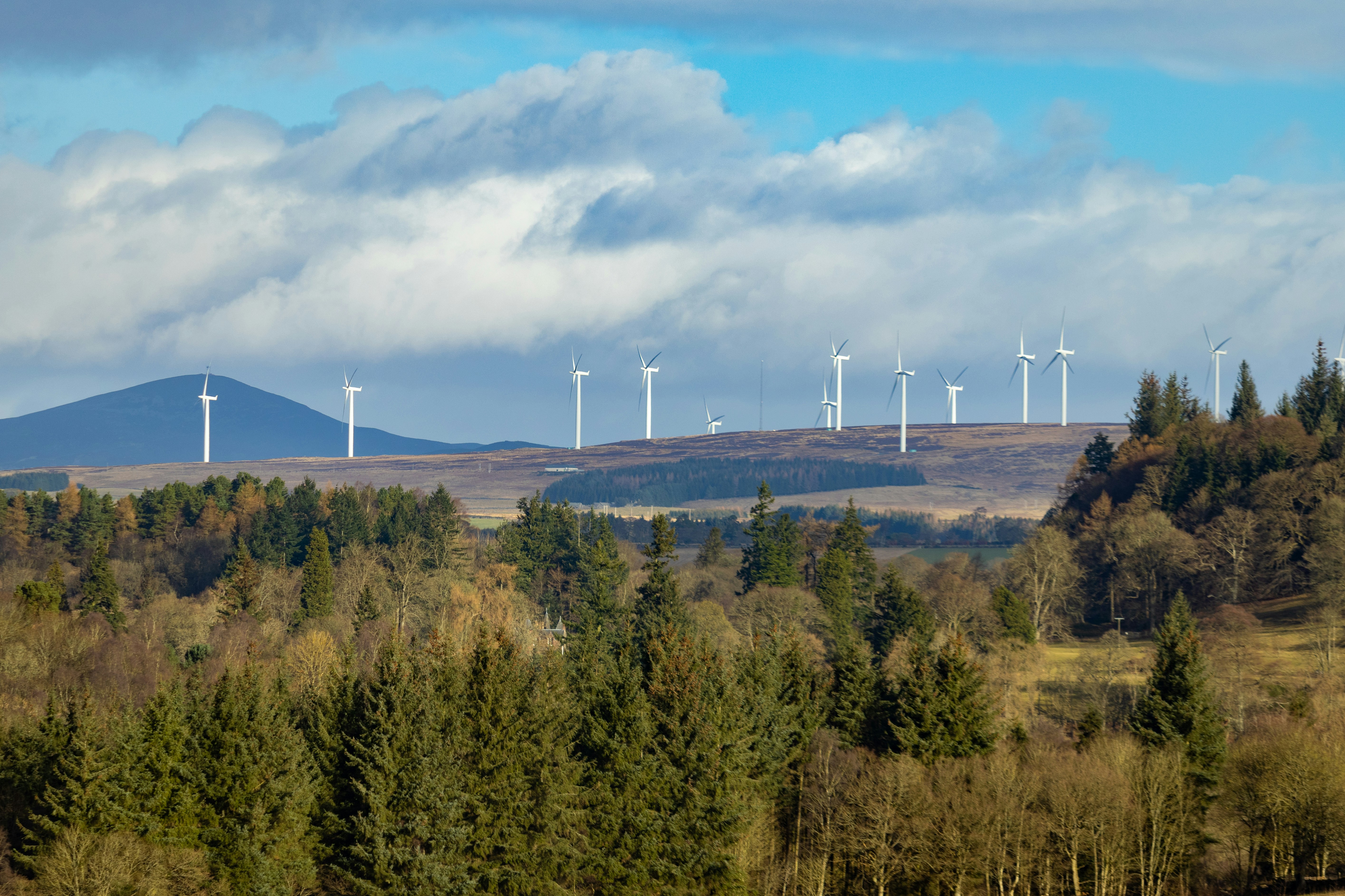 Onshore wind turbines stand across a rolling Scottish hillside above a dense forest, with mountains in the background and dramatic clouds stretching across a blue sky.