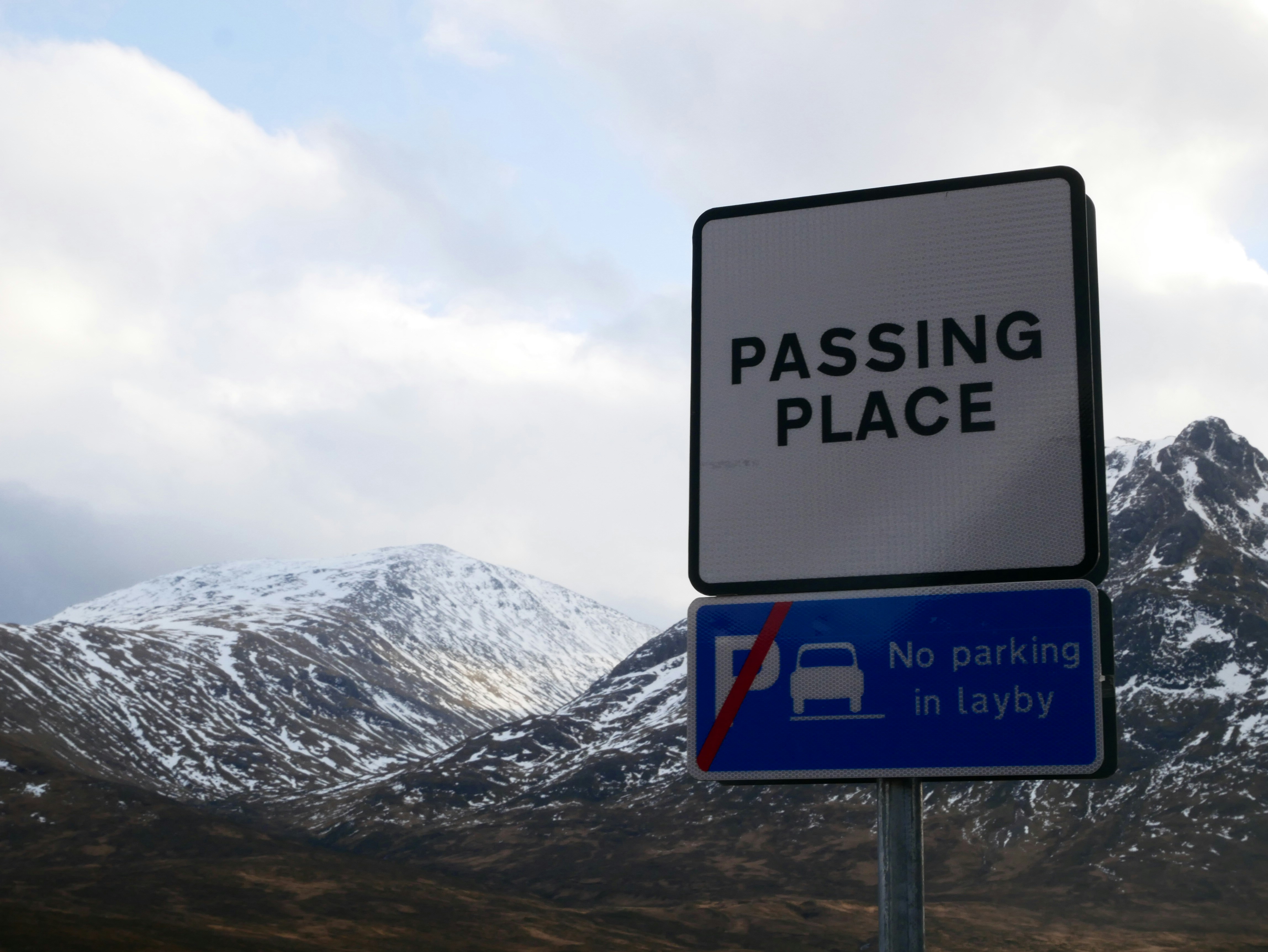 Roadside sign reading “PASSING PLACE” above another sign stating “No parking in layby,” set against a backdrop of snow-capped mountains and cloudy sky in a rural Scottish landscape.
