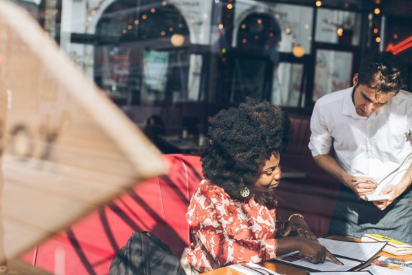Person seated at a café table reviewing a menu while a waiter stands beside the table, viewed through a café window with reflections.