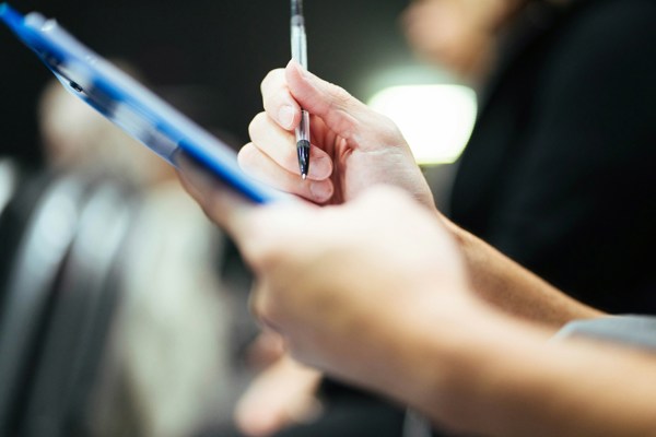 Close-up of a hand holding a pen and writing on a clipboard or form, with another blurred figure and document in the background, representing administrative or compliance tasks.