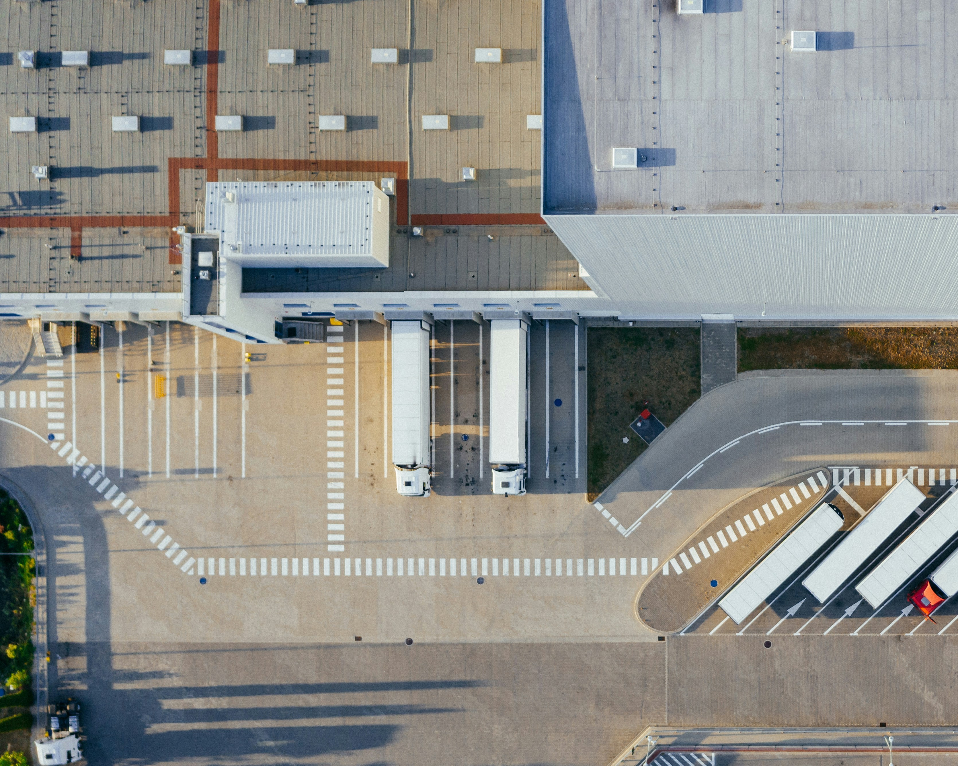 Aerial view of a modern industrial facility with loading docks and parked lorries. The image shows two white trucks positioned at loading bays, a large warehouse building with a grey roof, and marked lanes for vehicle movement.