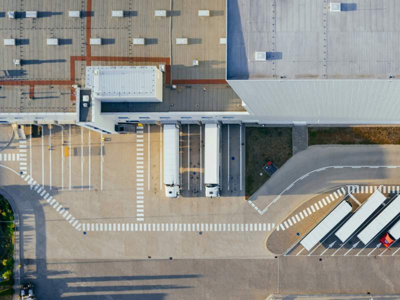 Aerial view of a modern industrial facility with loading docks and parked lorries. The image shows two white trucks positioned at loading bays, a large warehouse building with a grey roof, and marked lanes for vehicle movement.