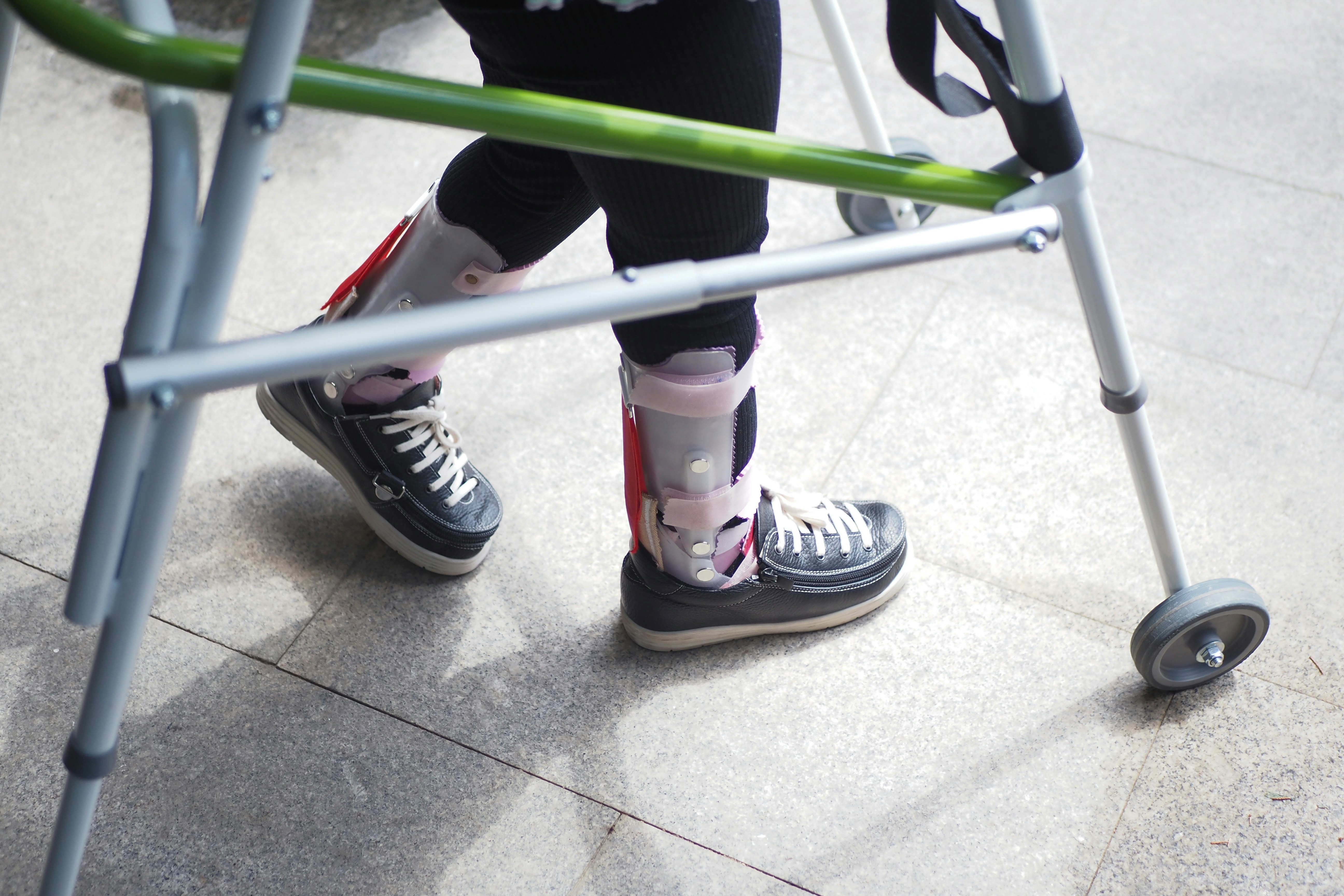 Close-up of a person using a metal walker for mobility, wearing black shoes and orthotic leg braces on both legs, walking on a tiled surface.