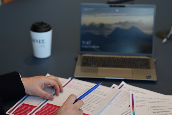 Person reviewing printed documents at a desk, with a pen in hand, and a laptop and coffee cup in the background.