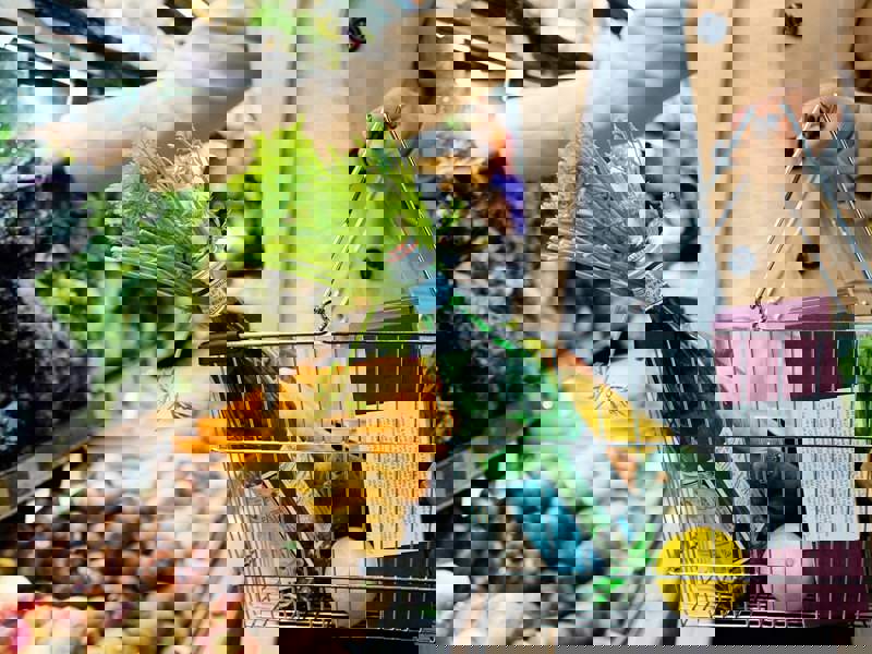 Person shopping in a grocery store, holding a metal basket filled with fresh produce including lemons, leafy greens, and bottled water, while selecting vegetables from a display of root crops and greens.