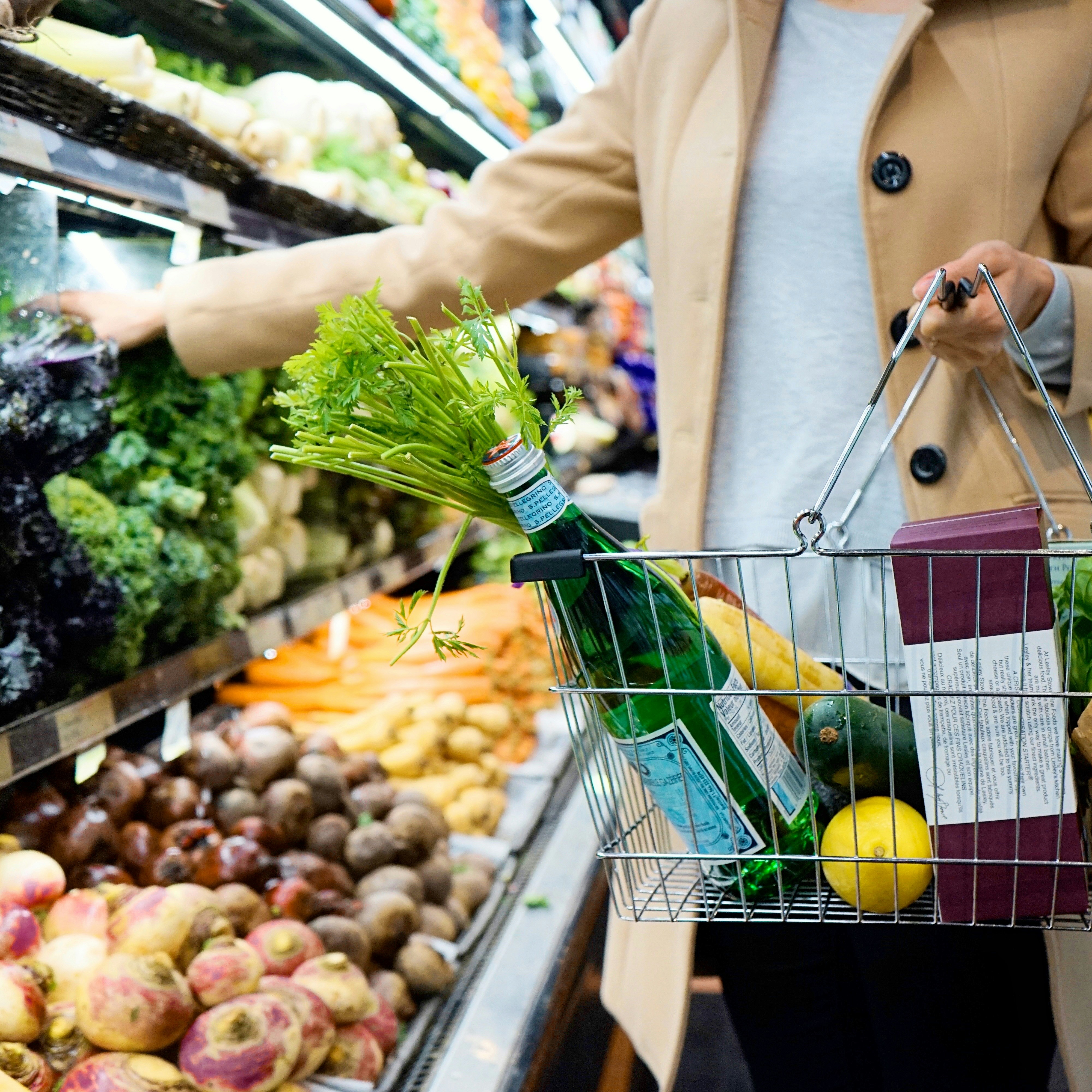 Person shopping in a grocery store, holding a metal basket filled with fresh produce including lemons, leafy greens, and bottled water, while selecting vegetables from a display of root crops and greens.