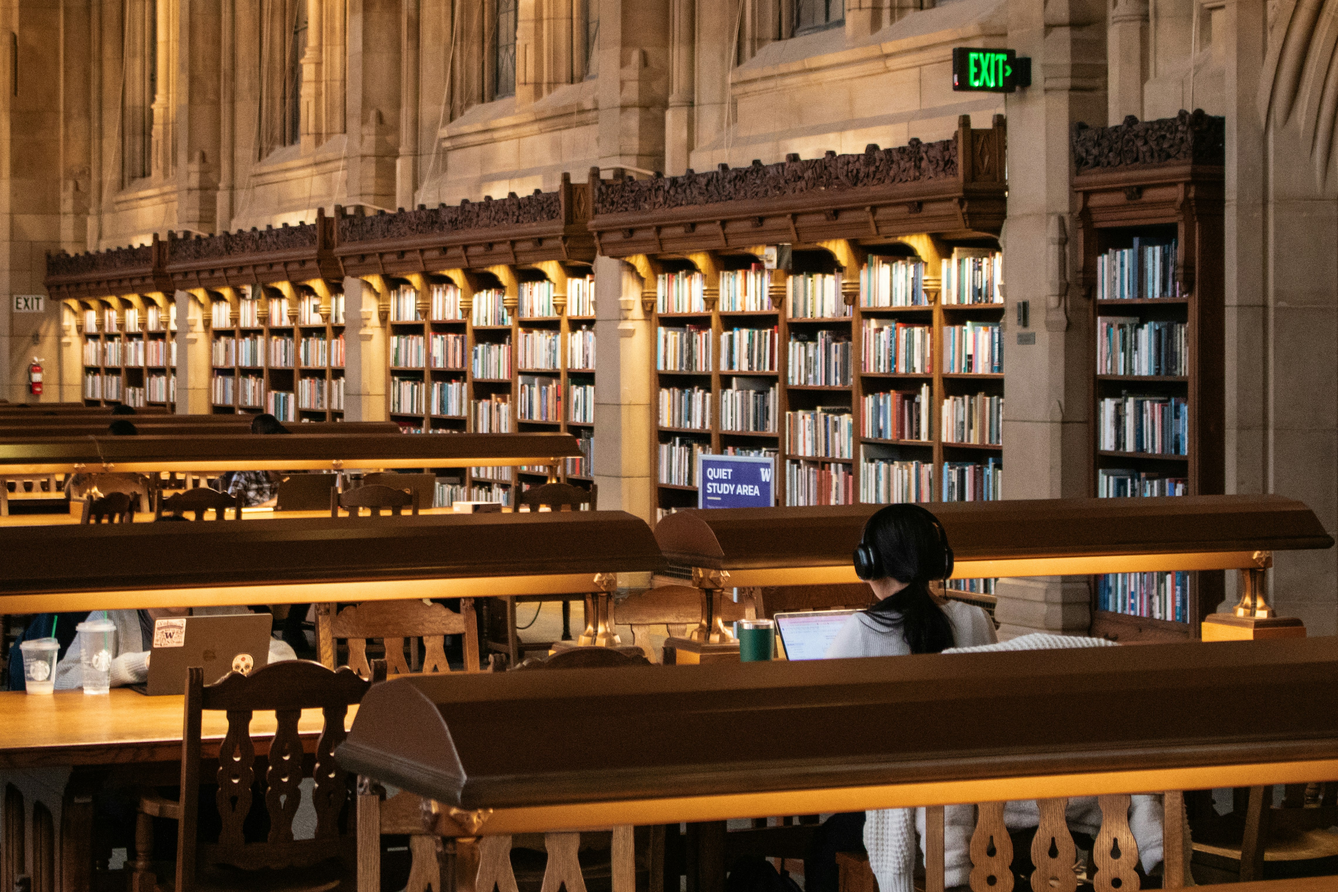 People seated at desks in a large library, studying and working among shelves of books in a formal academic setting.