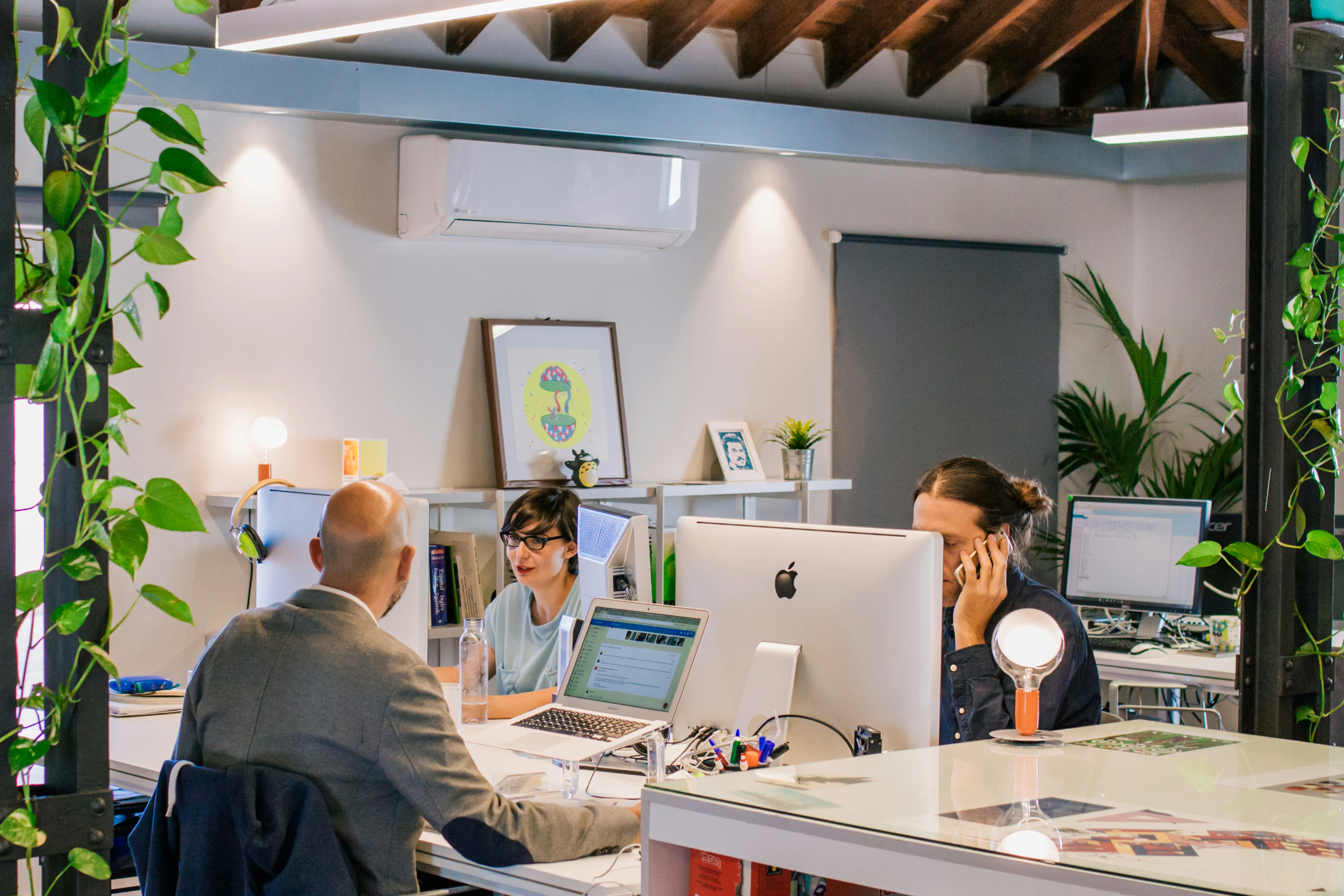 Office employees working collaboratively in a modern open-plan workplace with computers, desks and meeting spaces surrounded by indoor plants. The image represents workplace culture, employee engagement and organisational planning.