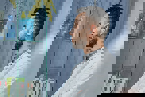 Pharmacist standing in a pharmacy, wearing a white coat and looking towards shelves of healthcare products.