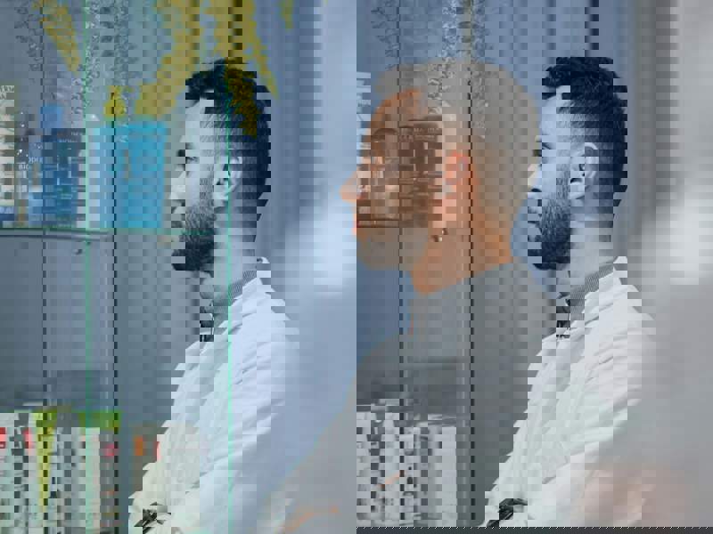 Pharmacist standing in a pharmacy, wearing a white coat and looking towards shelves of healthcare products.