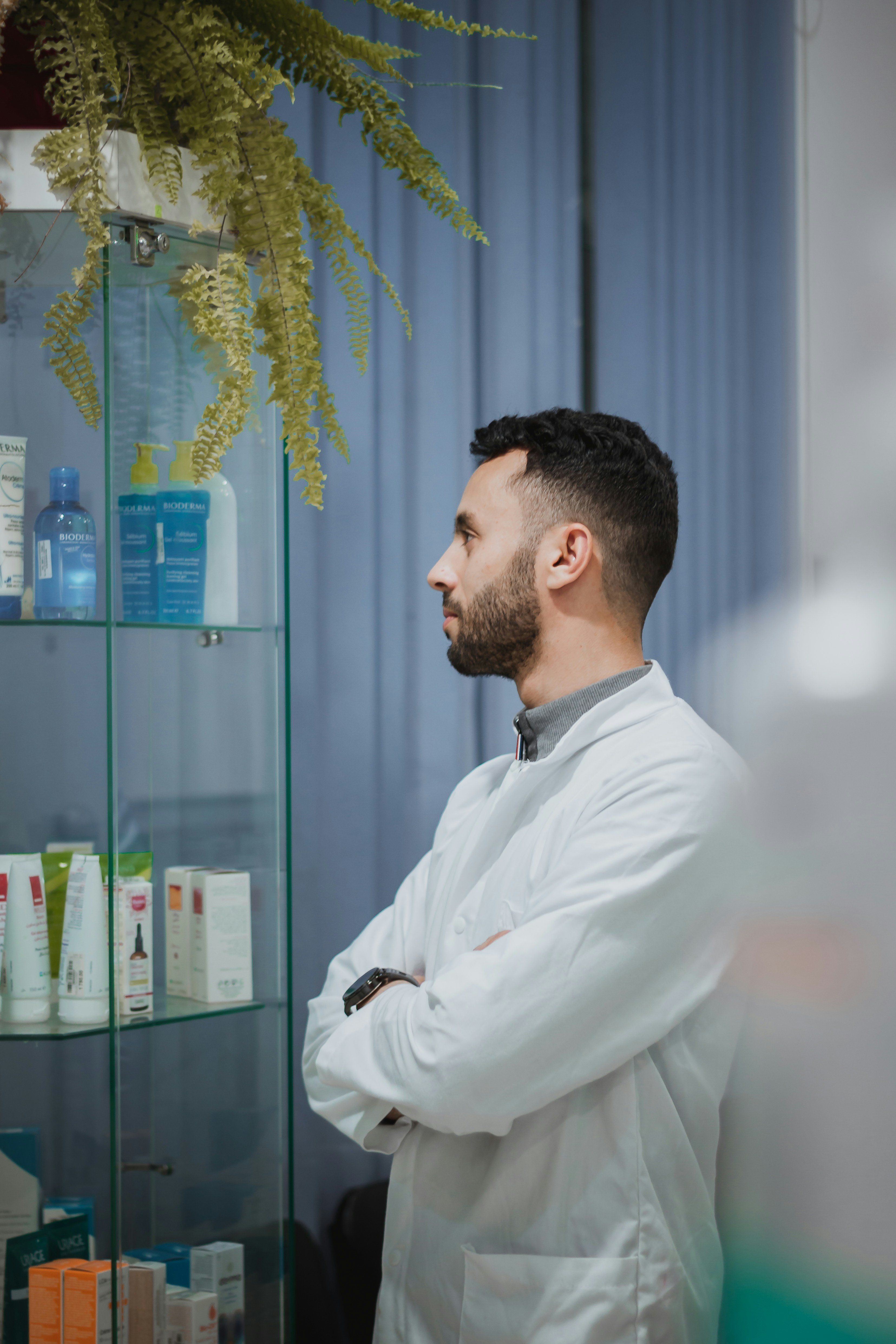 Pharmacist standing in a pharmacy, wearing a white coat and looking towards shelves of healthcare products.