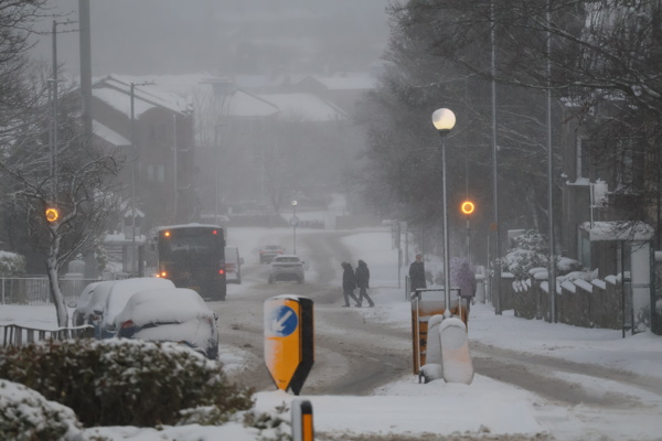 A snowy Aberdeen street scene with heavy snow covering the road, parked cars, and trees. A bus and a car are driving along the slushy road, while several pedestrians cross the street. Street lamps and traffic signs are visible, and the background shows houses partially obscured by falling snow.
