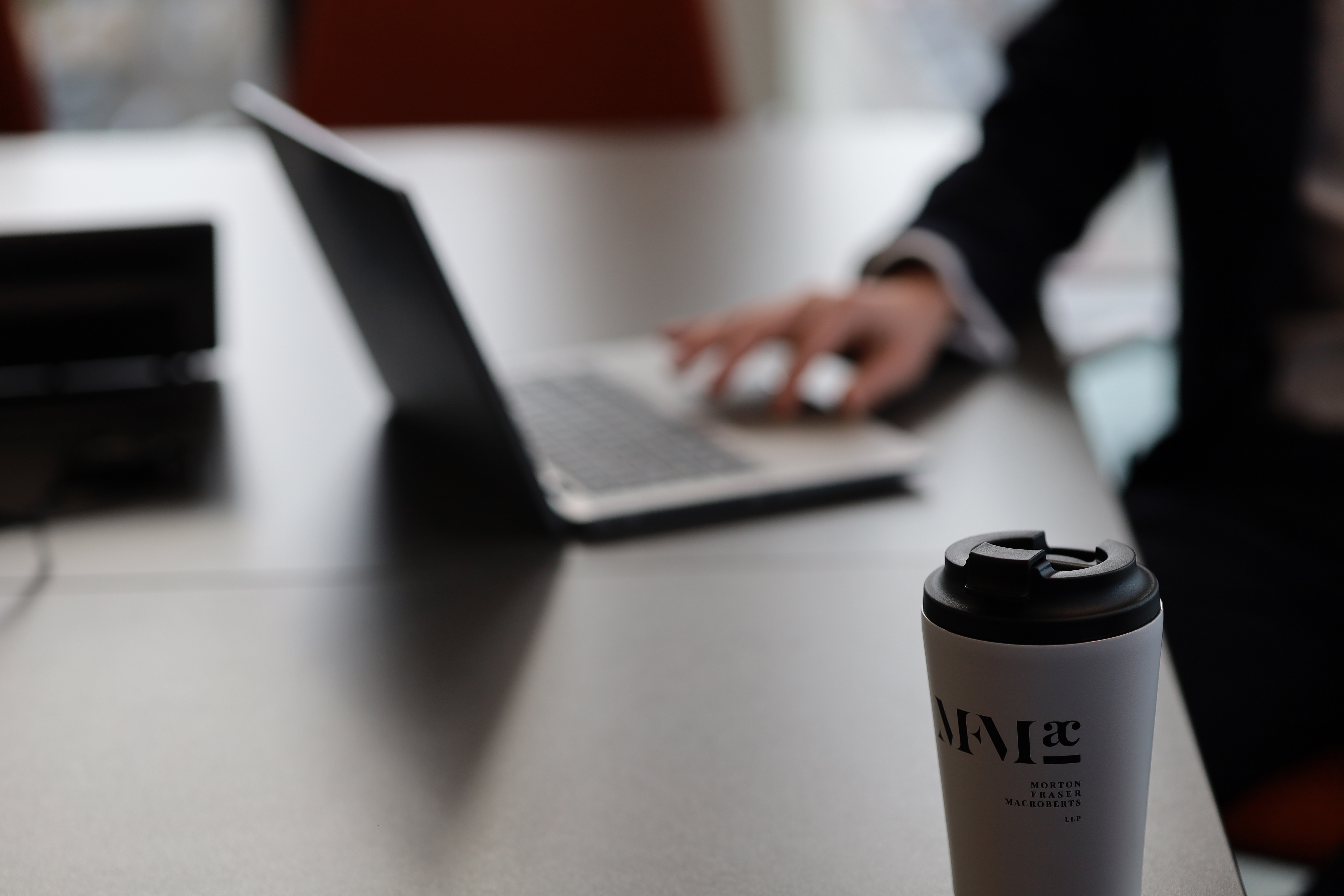Reusable coffee cup on an office desk with a blurred laptop and person working in the background
