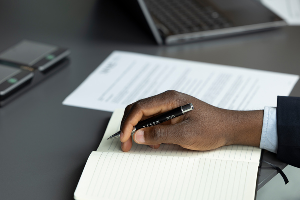 Hand writing notes in a notebook beside printed documents on a desk.