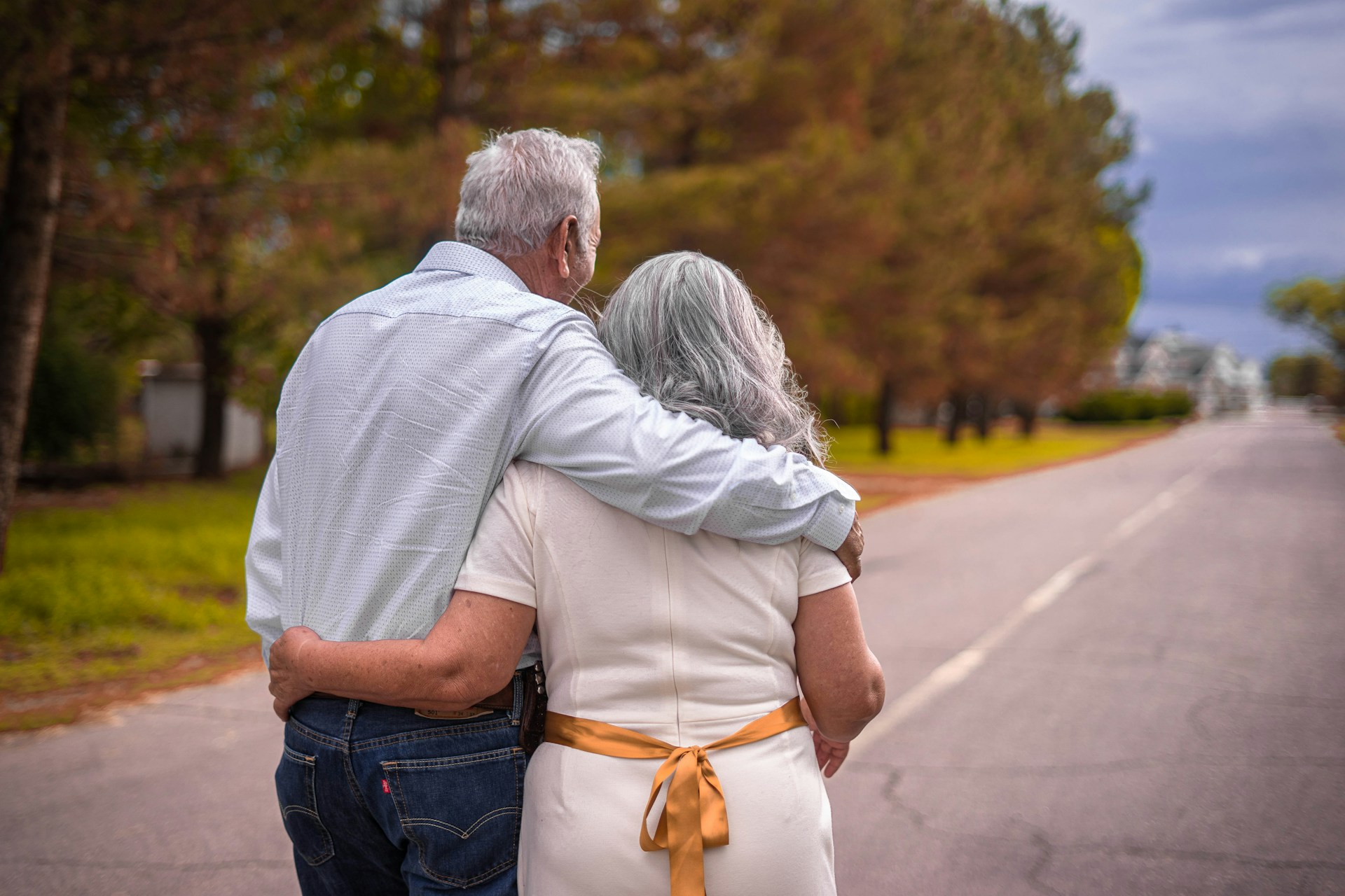 Two older adults walk arm in arm along a quiet tree-lined road, viewed from behind, with autumn-coloured trees and an open road stretching into the distance.
