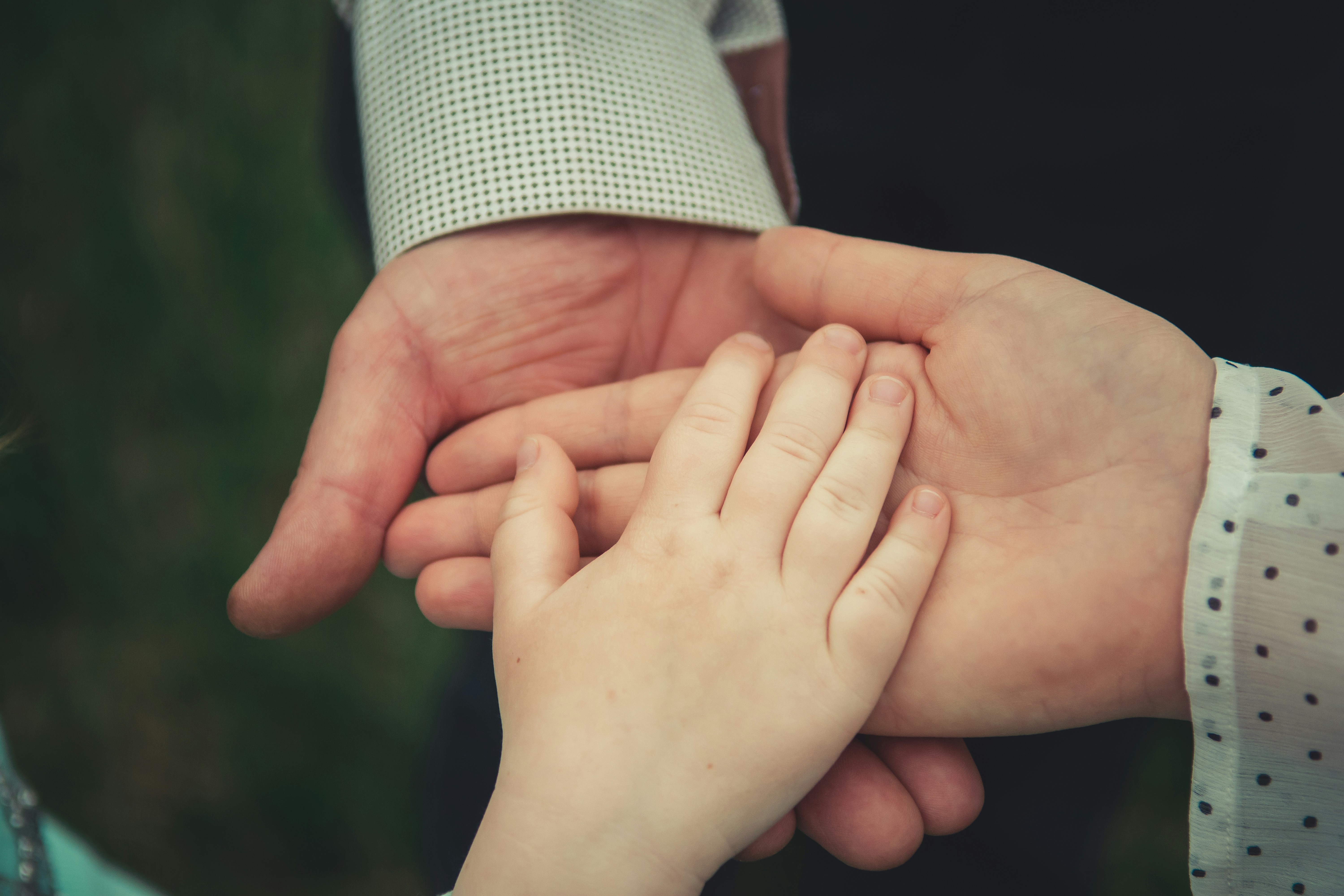 Two adult hands held palm up with a child's hand resting gently on one of them. The adults are wearing patterned long-sleeved shirts—one with small black dots and the other with a grid-like design.