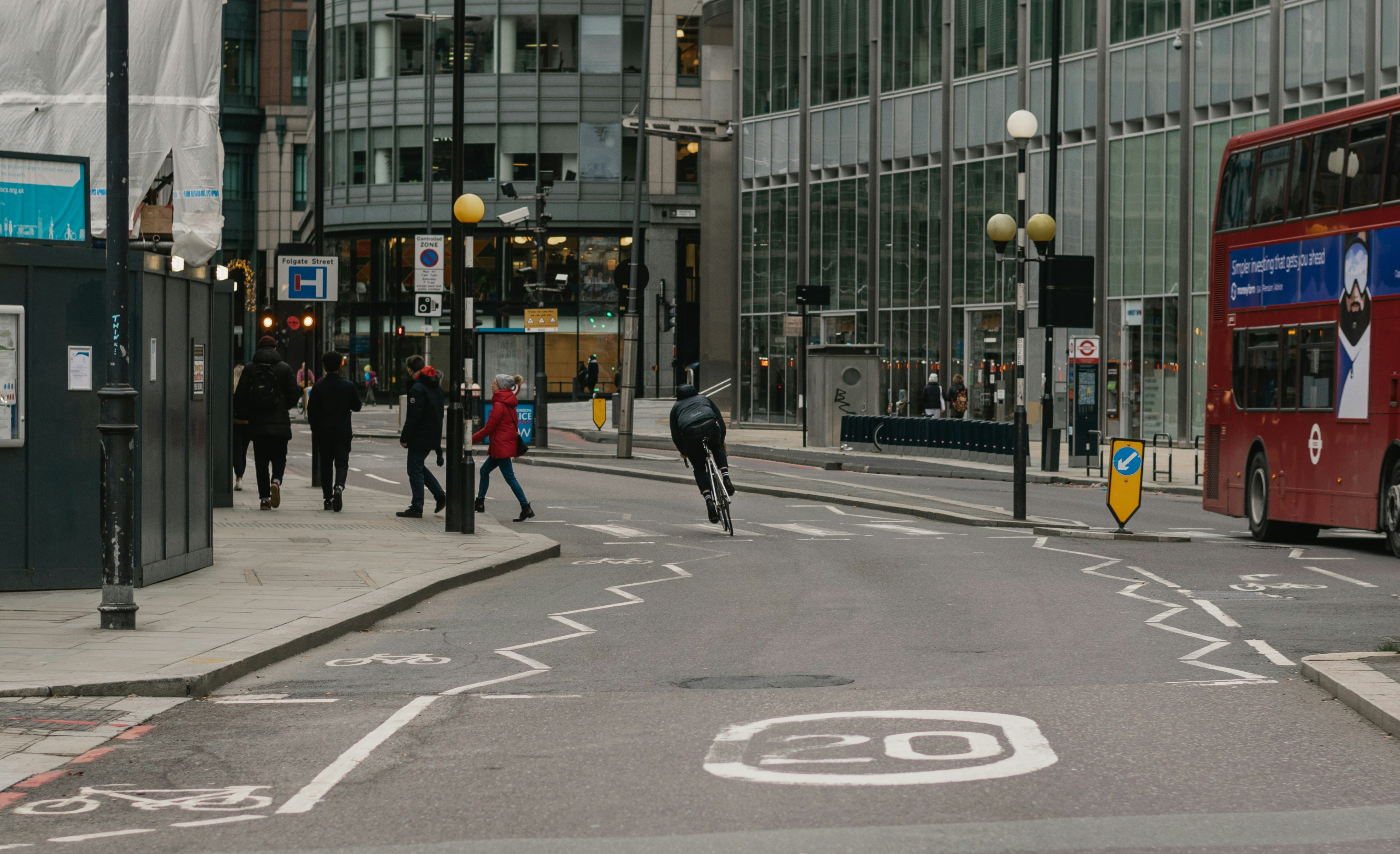 Cyclist riding through a city street junction with pedestrians crossing and a red double-decker bus beside modern office buildings.