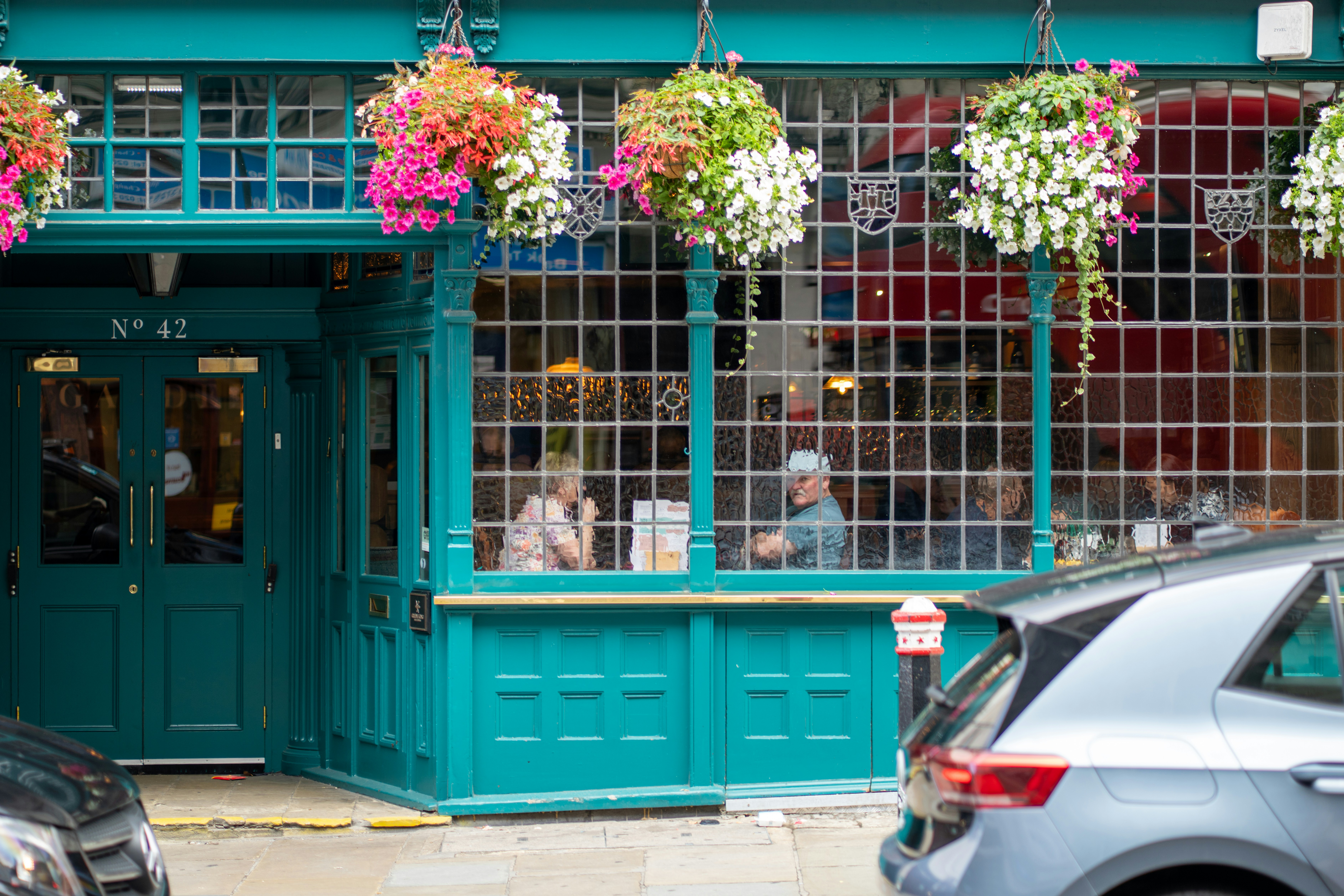 Exterior of a café with large windows, hanging flower baskets and people seated inside, viewed from the street.